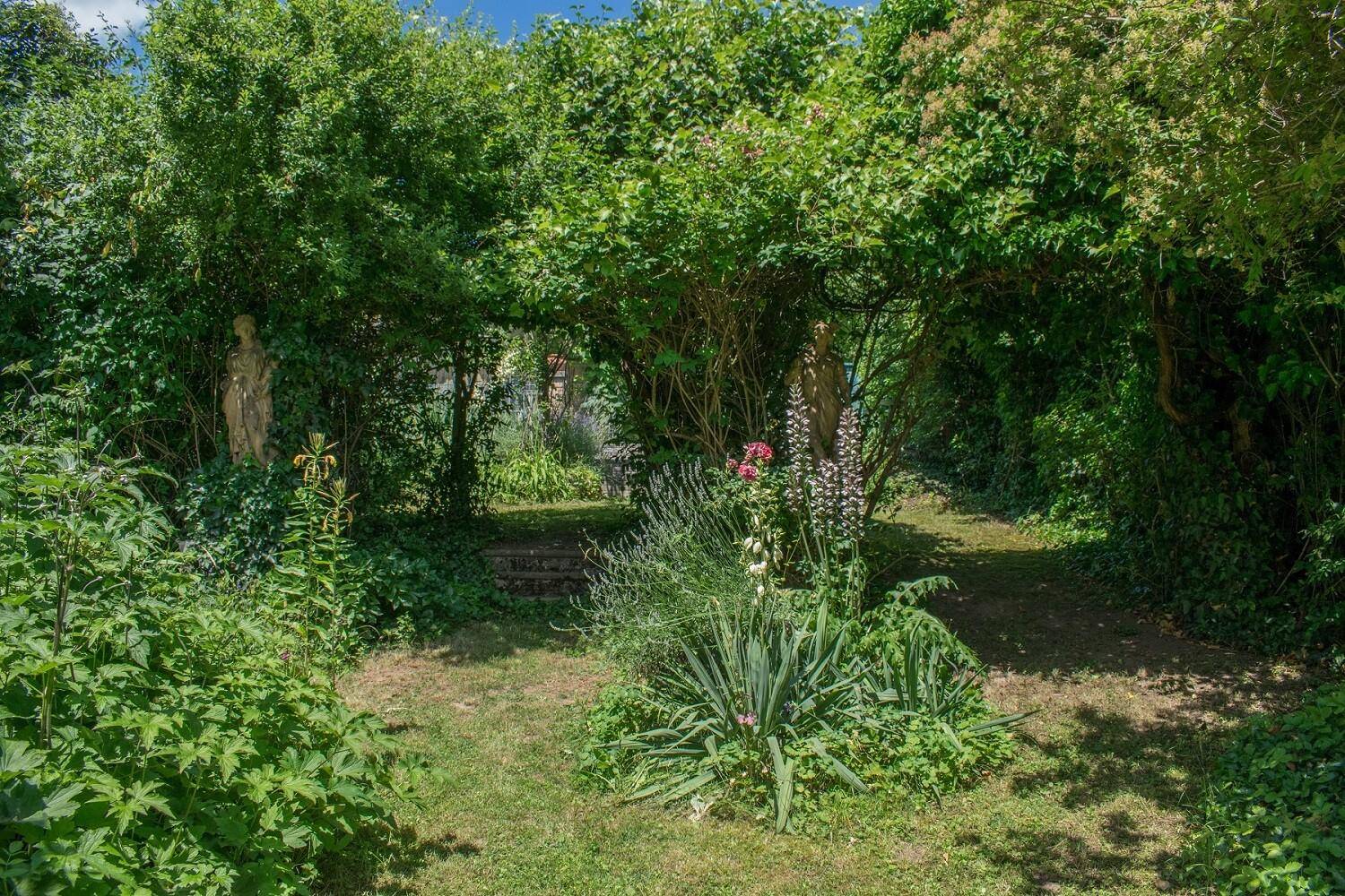 Verwunschener Gartenweg unter einem Blätterdach, gesäumt von üppigen Pflanzen und Statuen.