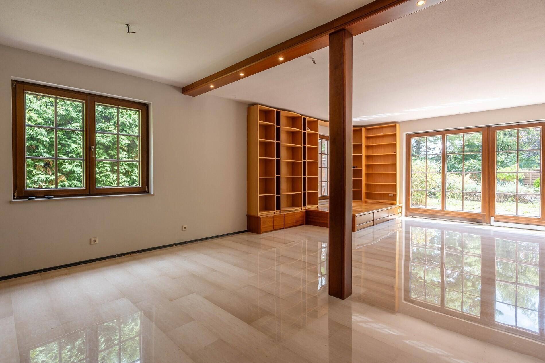 Helles Wohnzimmer mit glänzendem Fliesenboden, großen Fenstern und eingebauten Holzregalen.