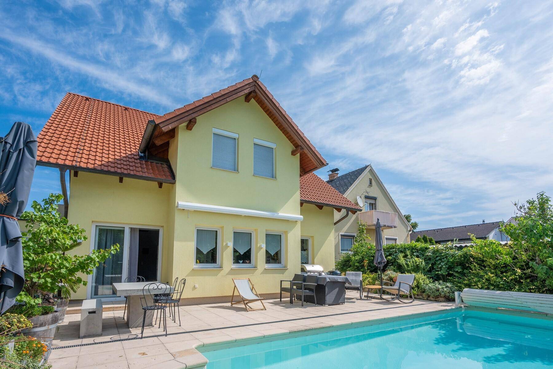 Sonniger Außenbereich mit großem Swimmingpool, Terrasse und Gartenmöbeln am Haus.