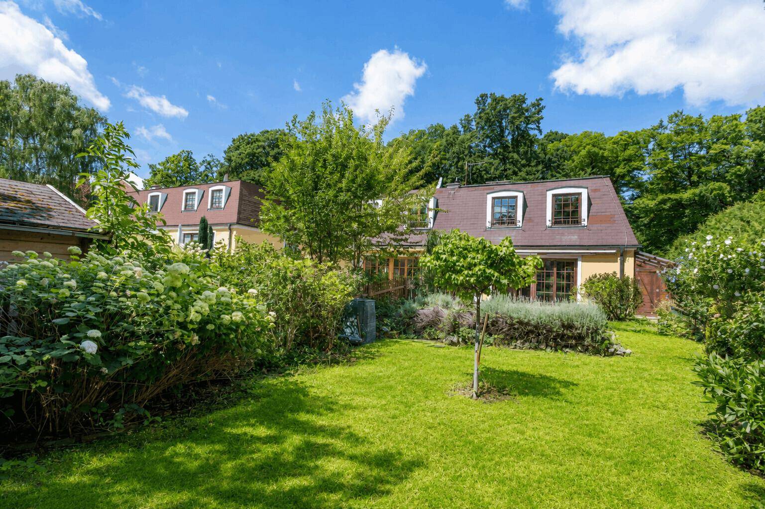 Ansicht des gelben Hauses mit Mansarddach vom grünen, sonnigen Garten aus.