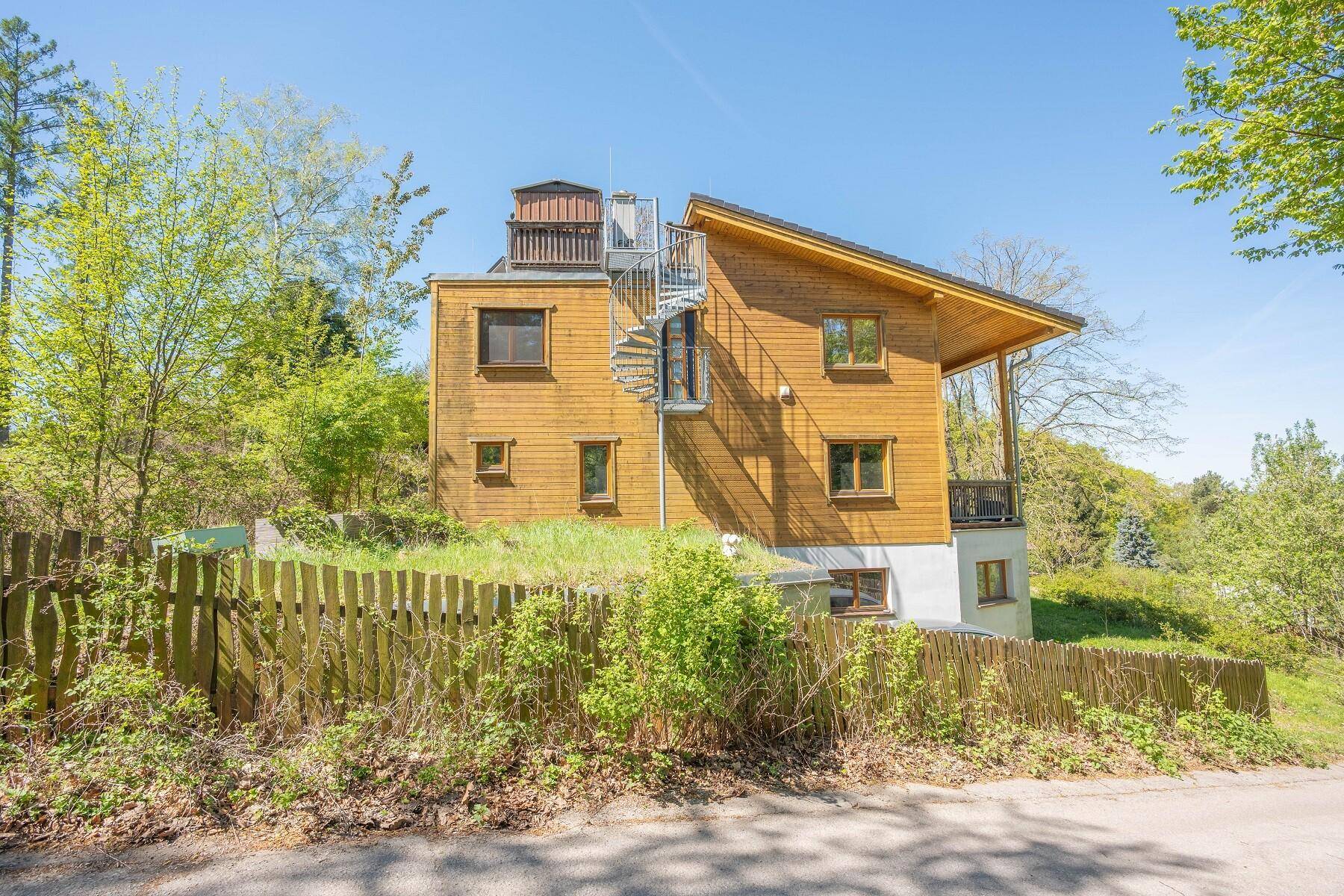 Ansicht des Holzhauses von der Seite mit einer Wendeltreppe und umgebender Natur.