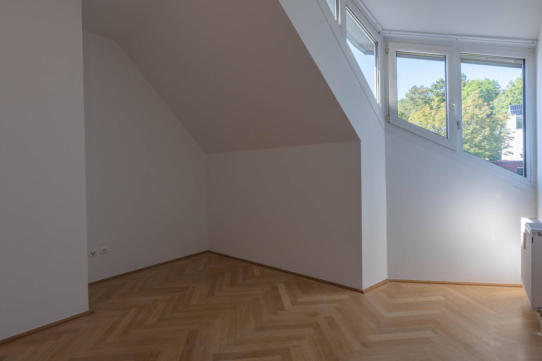 Helles Zimmer mit schrägen Wänden, Fischgrätparkett und großen Fenstern.