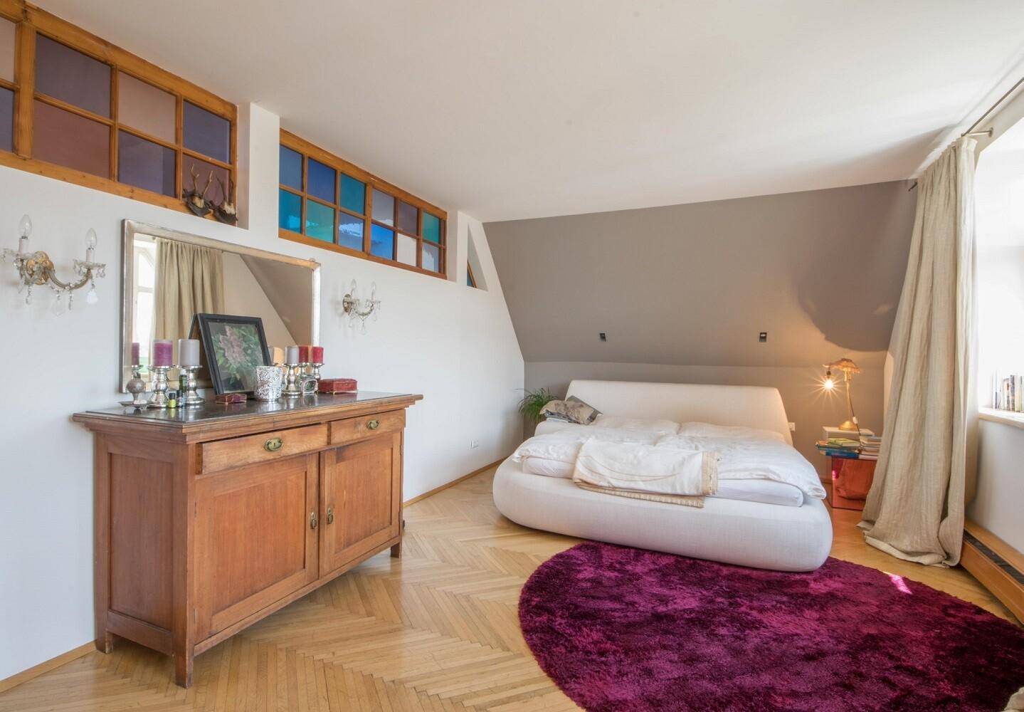 Schlafzimmer mit Dachschräge, Parkettboden und farbigen Fenstern, ausgestattet mit einem bequemen Bett.