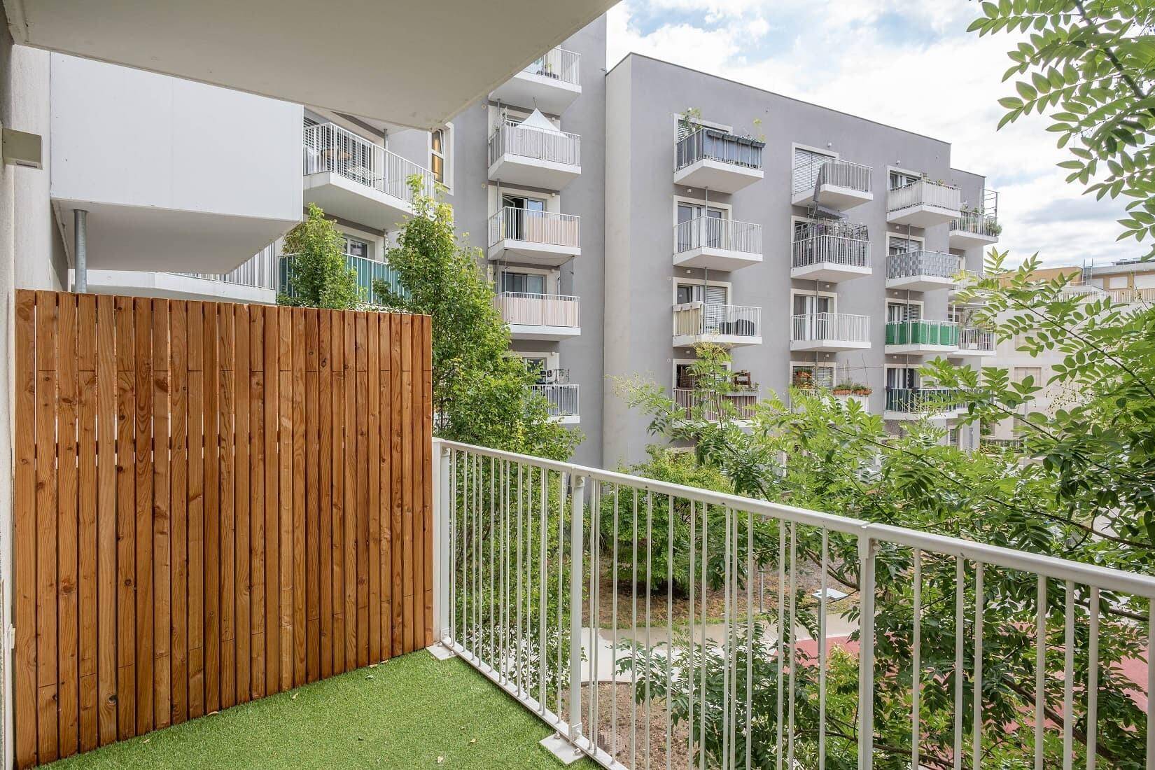 Balkon mit Kunstrasen und Holzsichtschutz, umgeben von Bäumen und modernen Wohngebäuden.