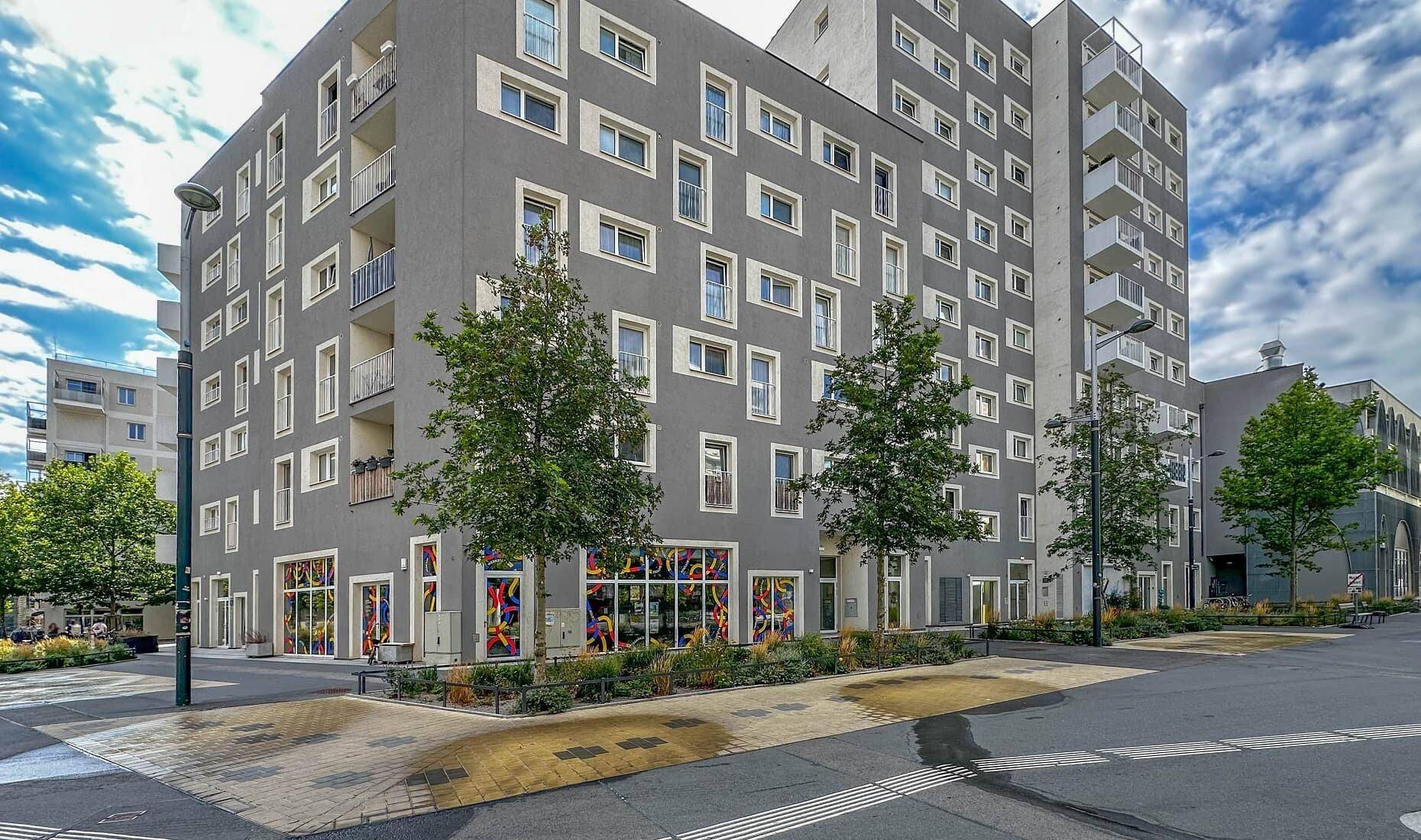Modernes Apartmentgebäude mit grauer Fassade, vielen Fenstern und Balkonen in urbaner Umgebung.
