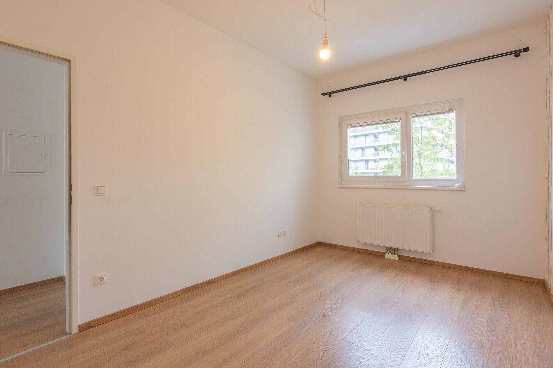 Helles, leeres Zimmer mit Holzboden und Fenster, das einen Blick auf umliegende Gebäude bietet.