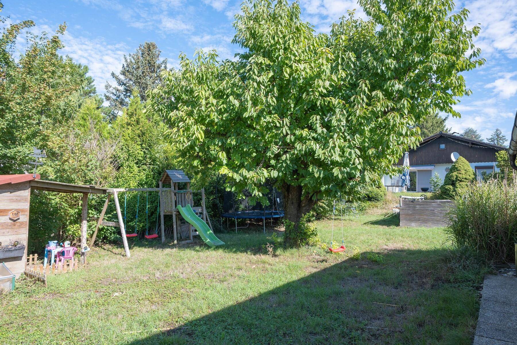 Weitläufiger Gartenbereich mit Schaukel, Rutsche und Gartenhaus unter einem großen Baum.