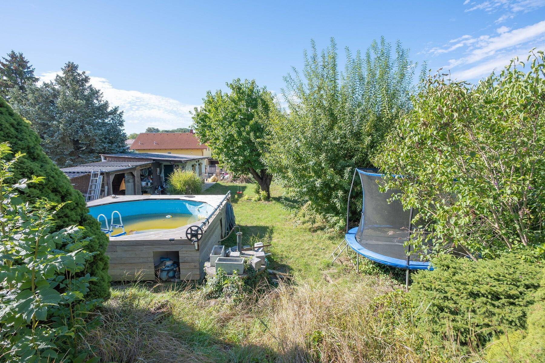 Gartenbereich mit Aufstellpool, Trampolin und viel Grünfläche unter blauem Himmel.