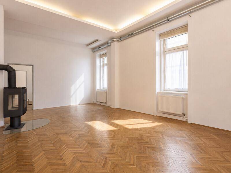 Unmöblierter Wohnbereich mit Fischgrätparkett, Kaminofen und viel Tageslicht durch große Fenster.