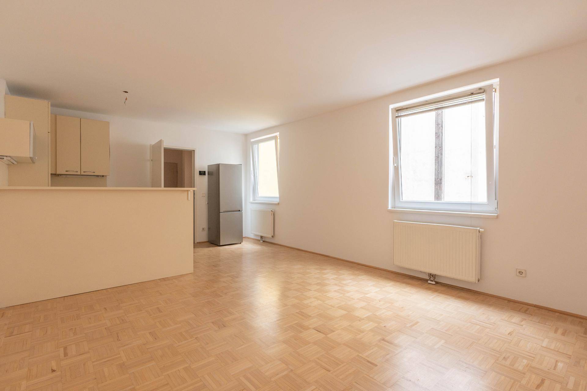 Heller Wohnbereich mit Parkettboden, zwei Fenstern und integrierter Küchenzeile mit Kühlschrank.
