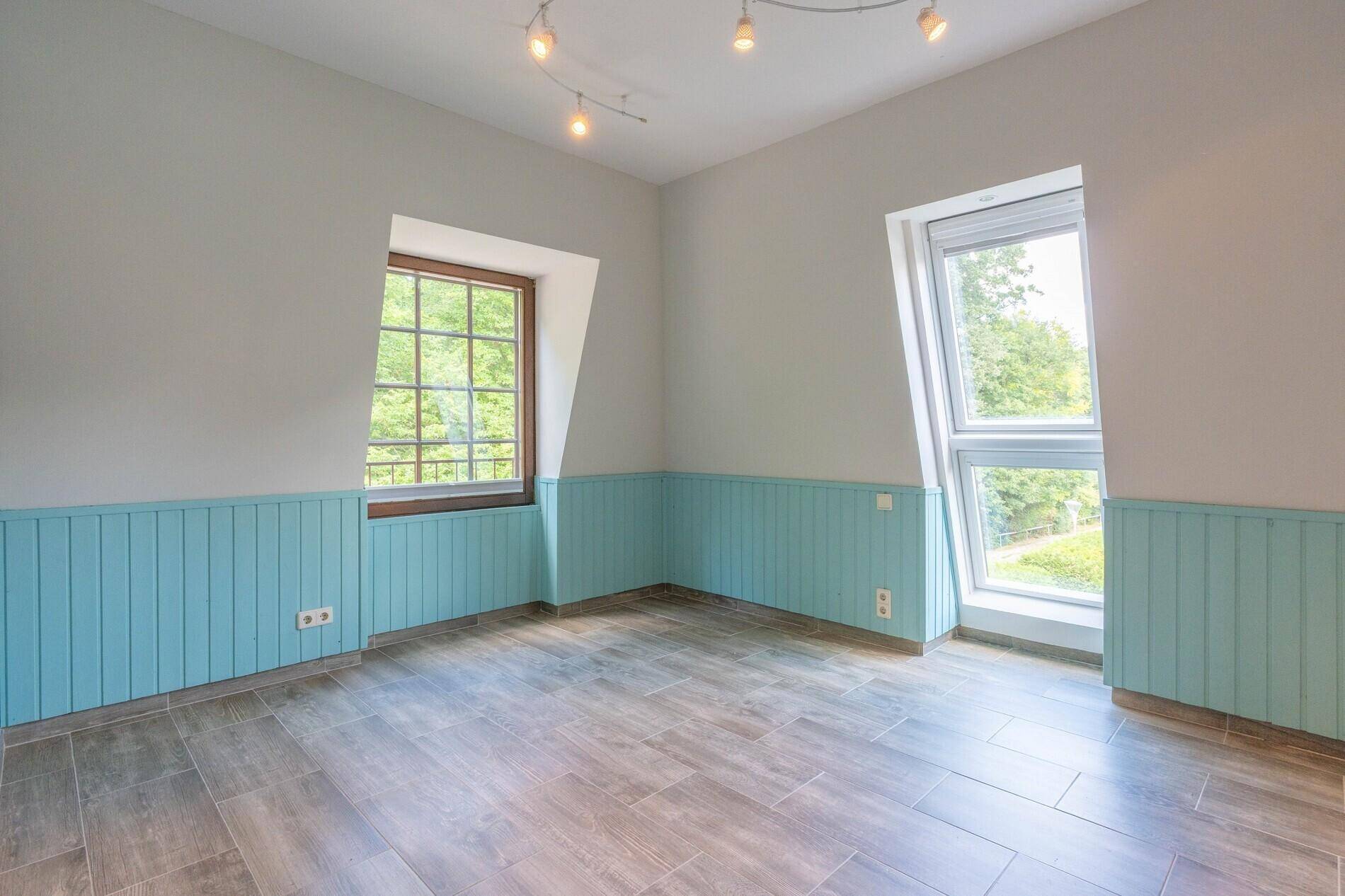 Helles Zimmer mit hellblauer Holzvertäfelung, zwei Fenstern und modernem Fliesenboden in Holzoptik.