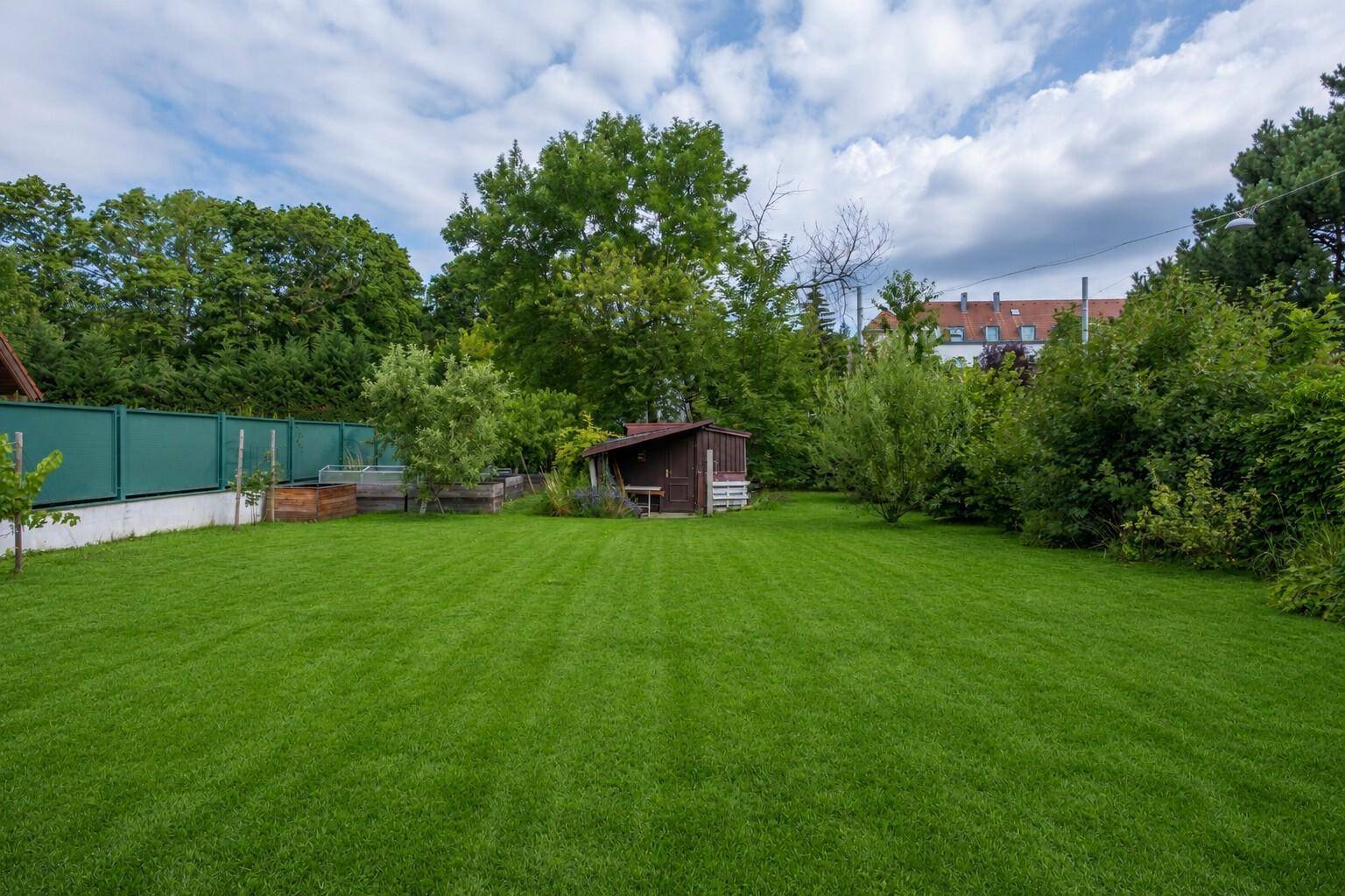 Große Rasenfläche im Garten mit einem kleinen Holzschuppen im Hintergrund, umgeben von Bäumen und Sträuchern.