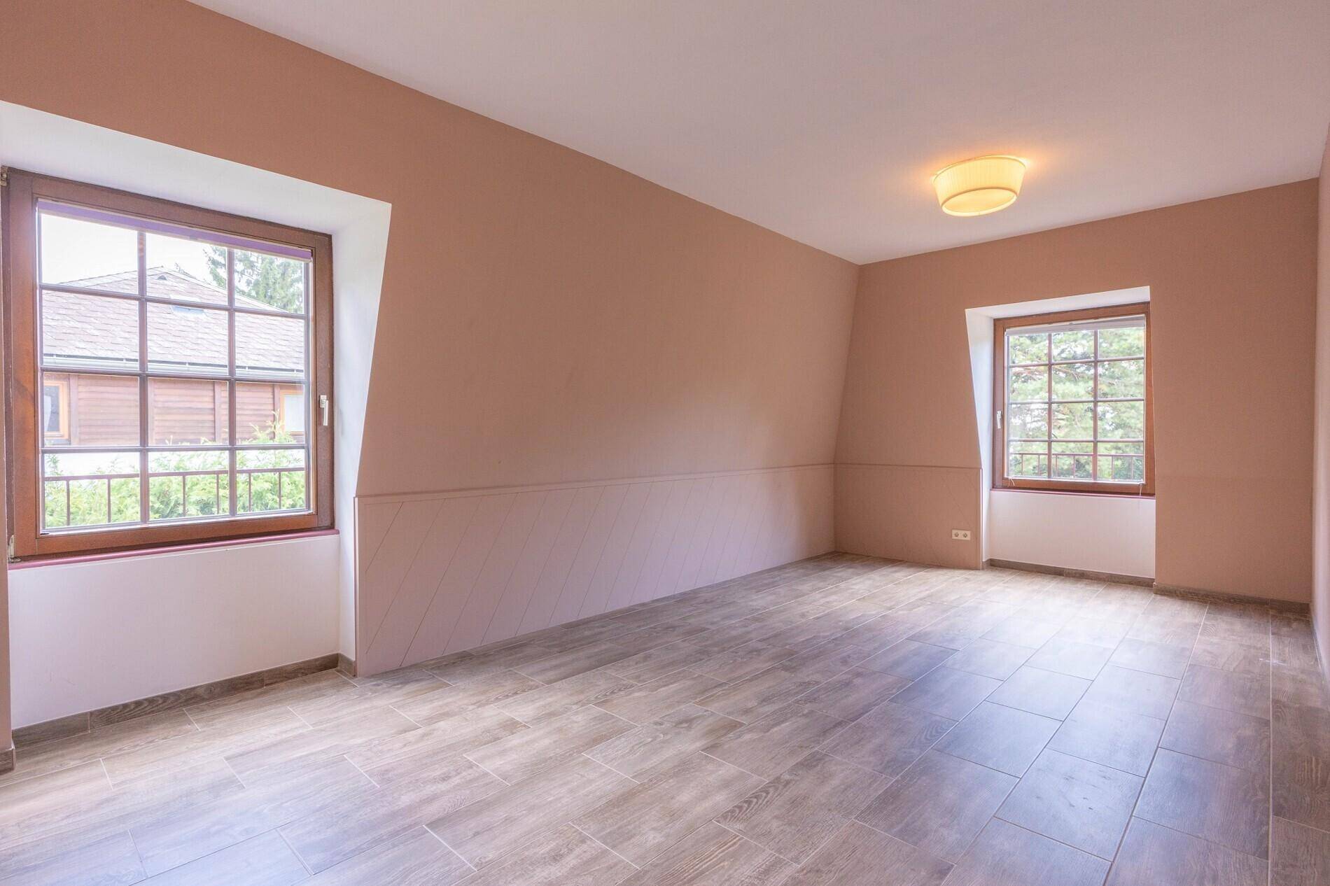Helles Schlafzimmer mit taupefarbenen Wänden, Fliesenboden in Holzoptik und zwei Fenstern.