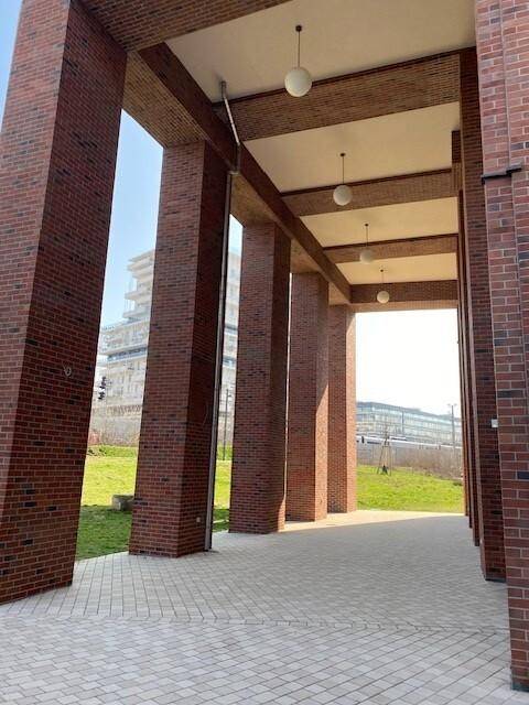 Überdachter Gang unter den Ziegelsäulen des Gebäudes, mit Blick auf eine Grünfläche und weitere moderne Bauten.