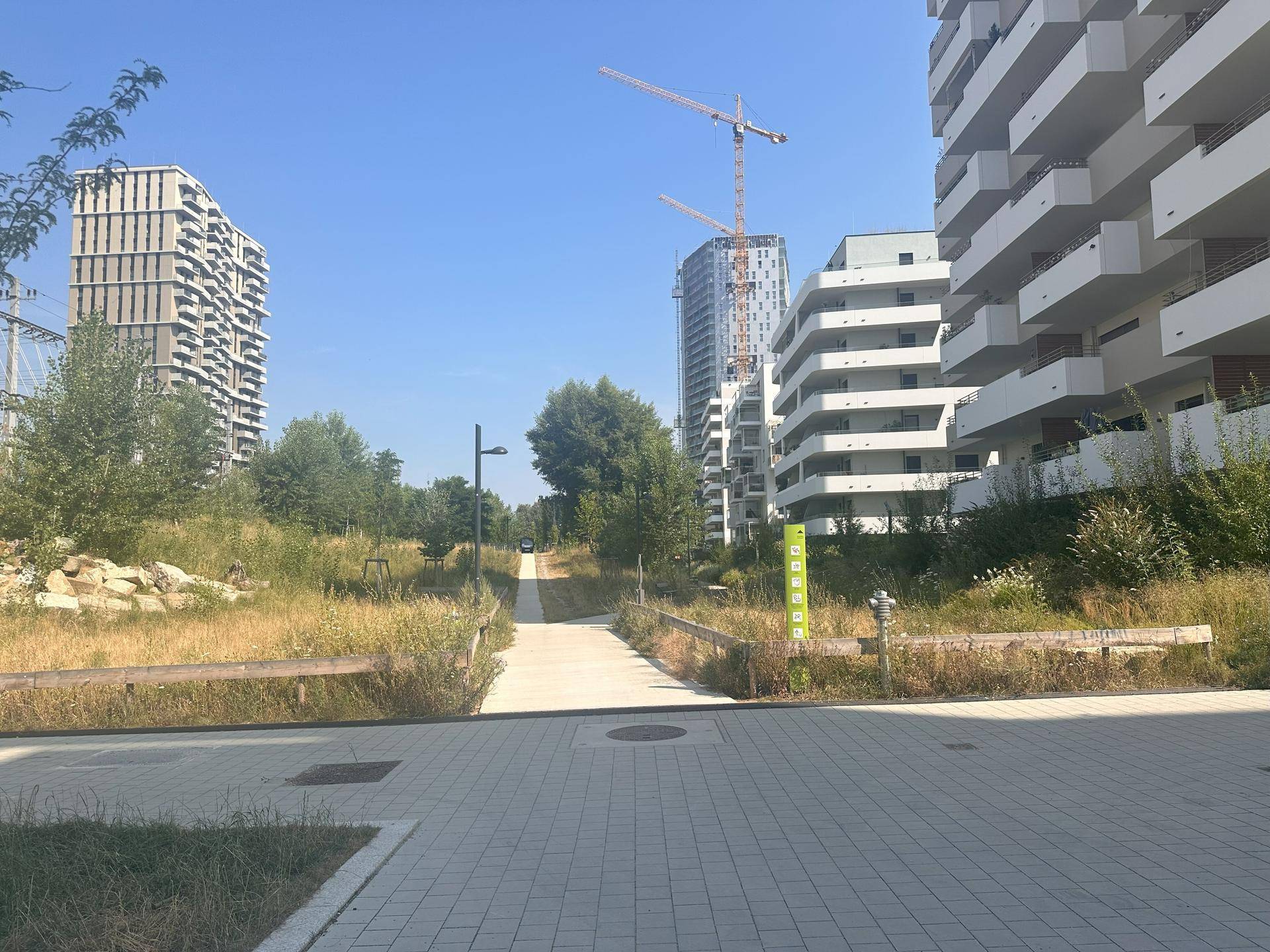 Weitläufige Straßenansicht mit modernen Wohngebäuden, einem grünen Weg und Baukränen am Horizont.