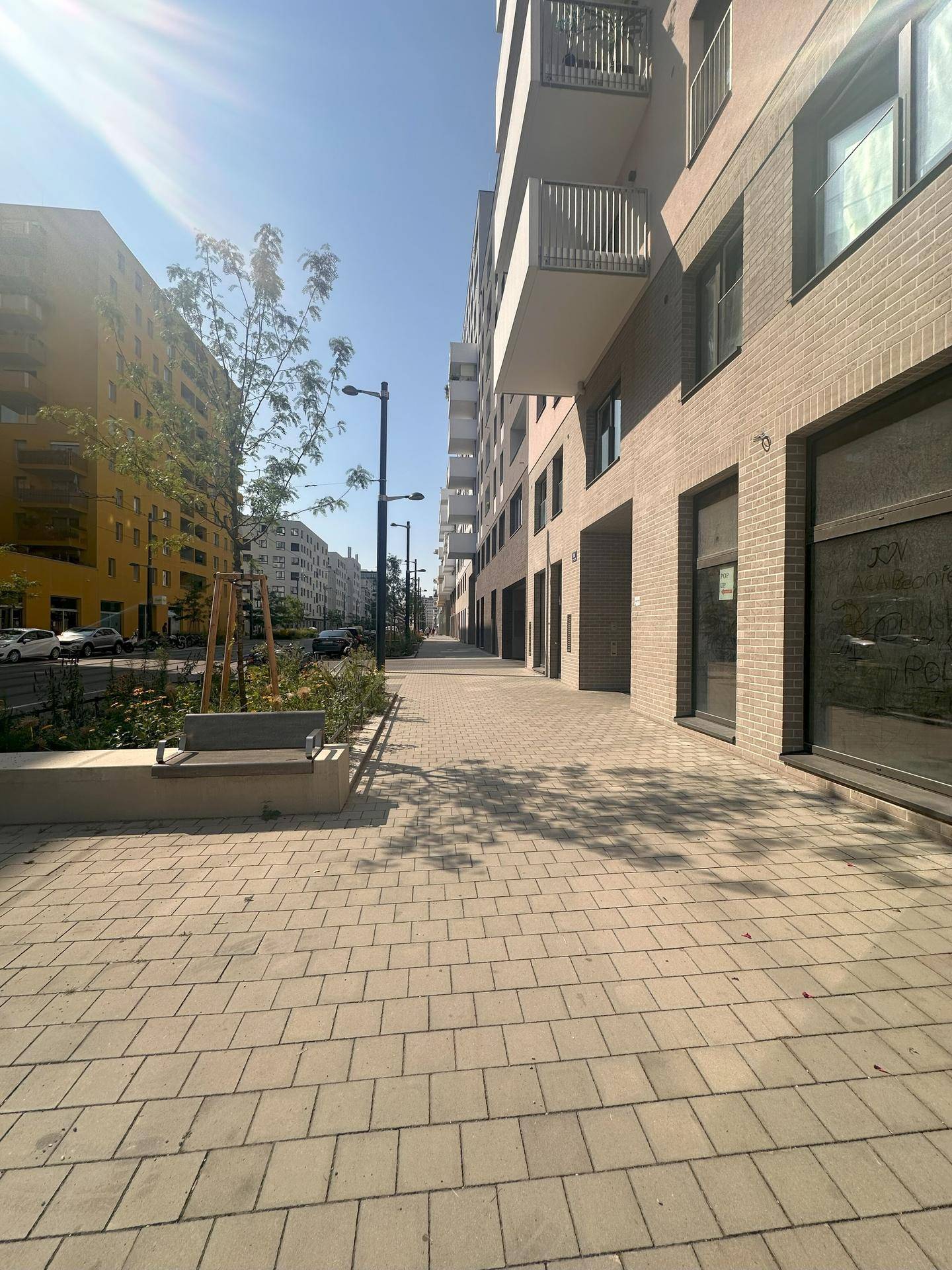 Langer Gehweg entlang eines modernen Gebäudes mit Ziegelfassade, gesäumt von Bäumen und einer Sitzbank.
