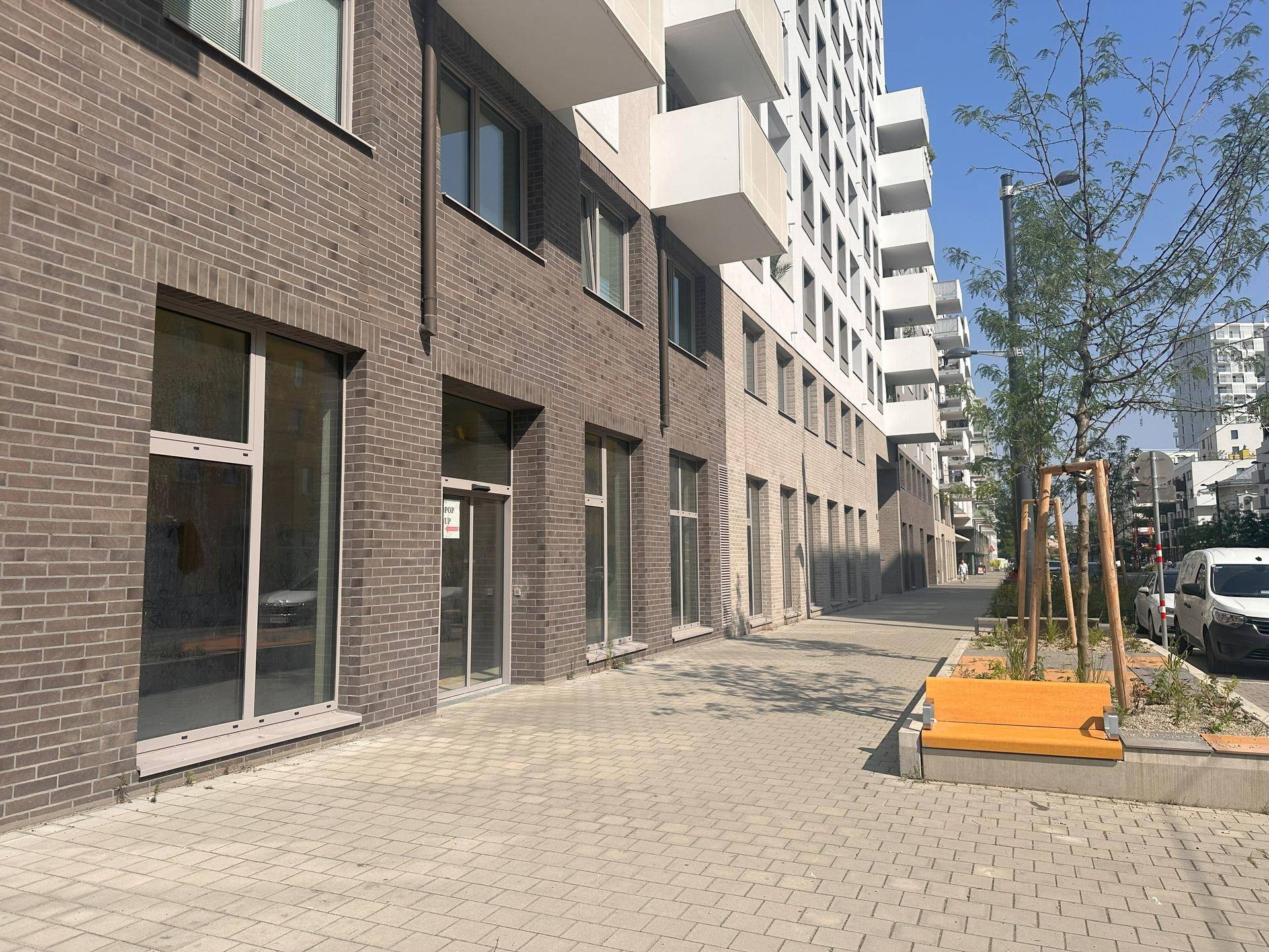 Gehweg entlang eines modernen Gebäudes mit Ziegelfassade im Erdgeschoss, großen Fenstern und einer Sitzbank.