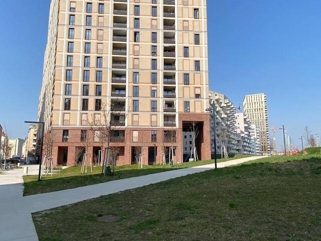 Hohes modernes Wohngebäude mit vielen Balkonen und einer Grünfläche im Vordergrund unter blauem Himmel.