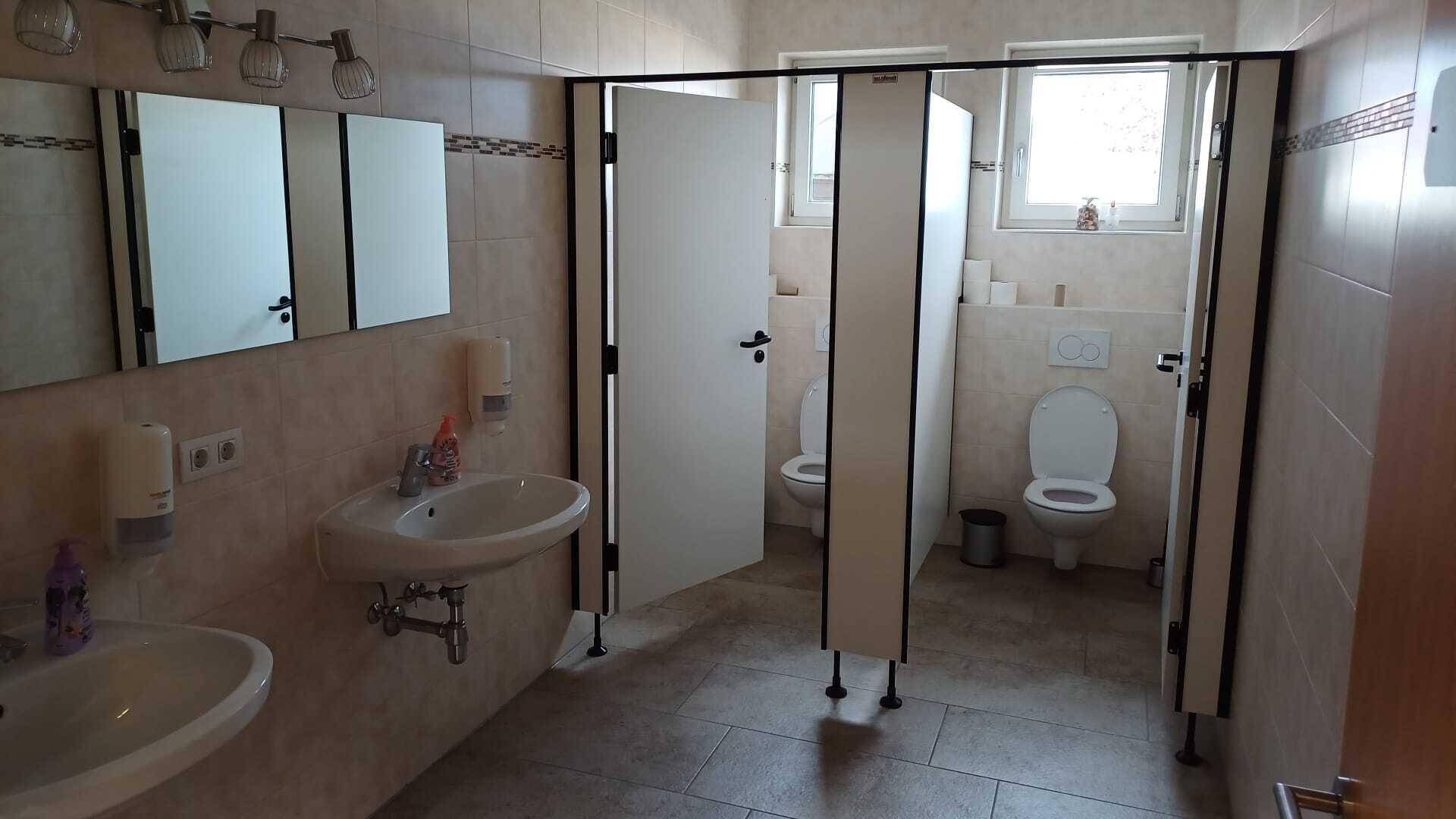 Geräumiger öffentlicher Toilettenbereich mit mehreren Kabinen, Waschbecken und Fenstern für Tageslicht.