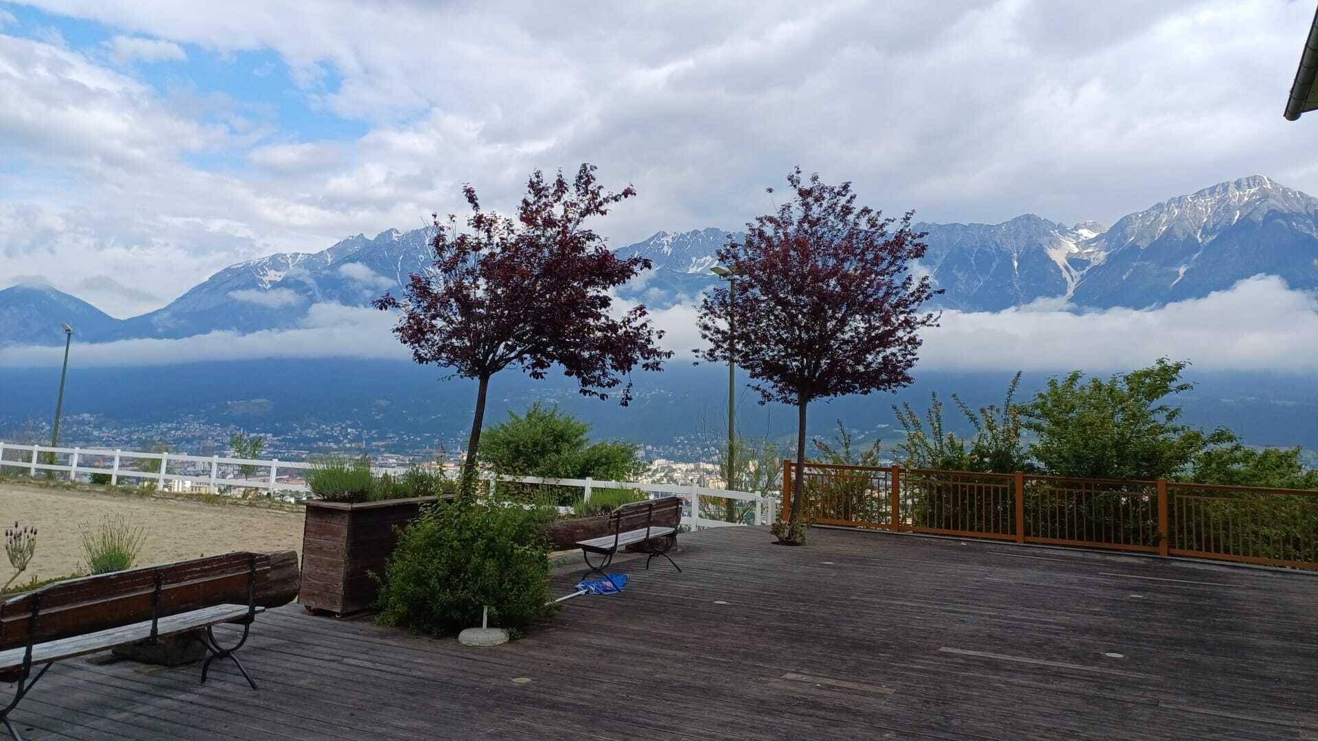 Holzterrasse mit Bänken und Bäumen, die einen weiten Blick auf die Berge und das Tal bietet.