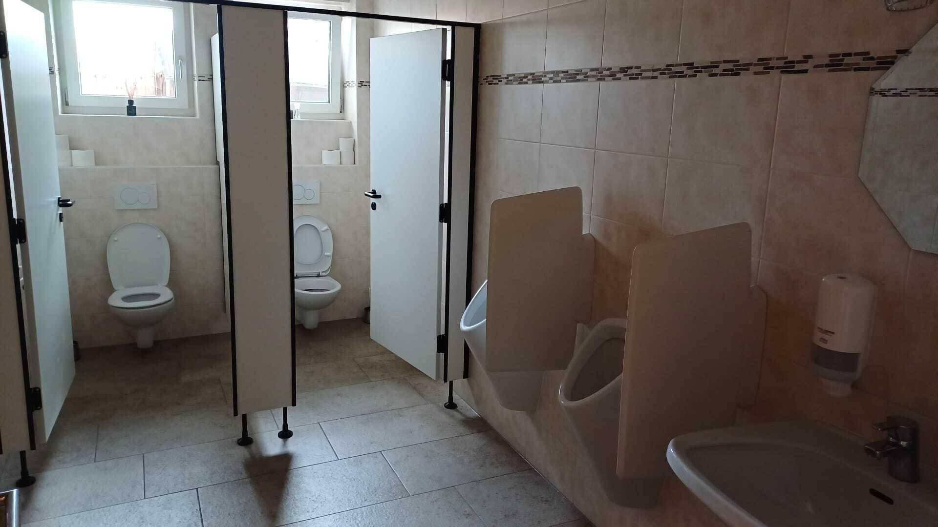 Öffentlicher Toilettenbereich mit mehreren Kabinen, Urinalen und Waschbecken, hell gefliest.