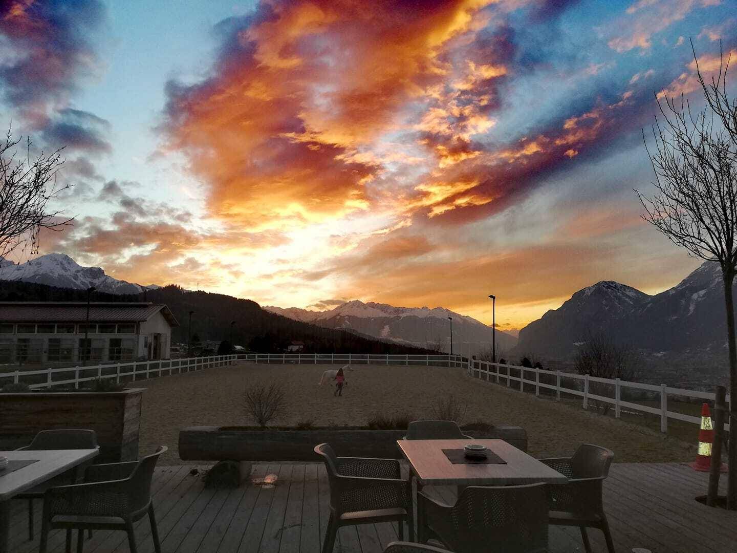 Terrasse mit Sitzgelegenheiten und Blick auf einen Reitplatz bei Sonnenuntergang über den Bergen.