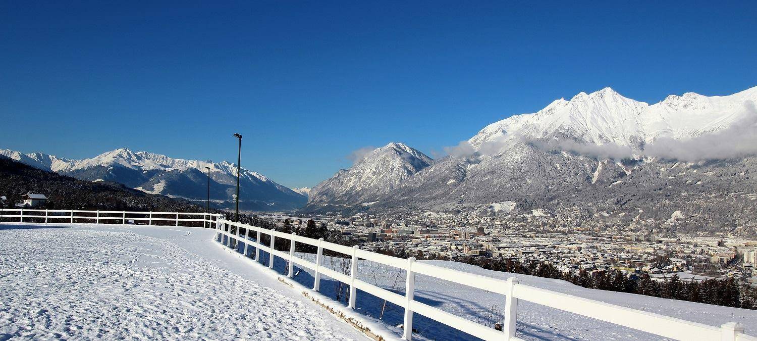 Winterlandschaft mit schneebedecktem Reitplatz und majestätischen Bergen im Hintergrund unter blauem Himmel.