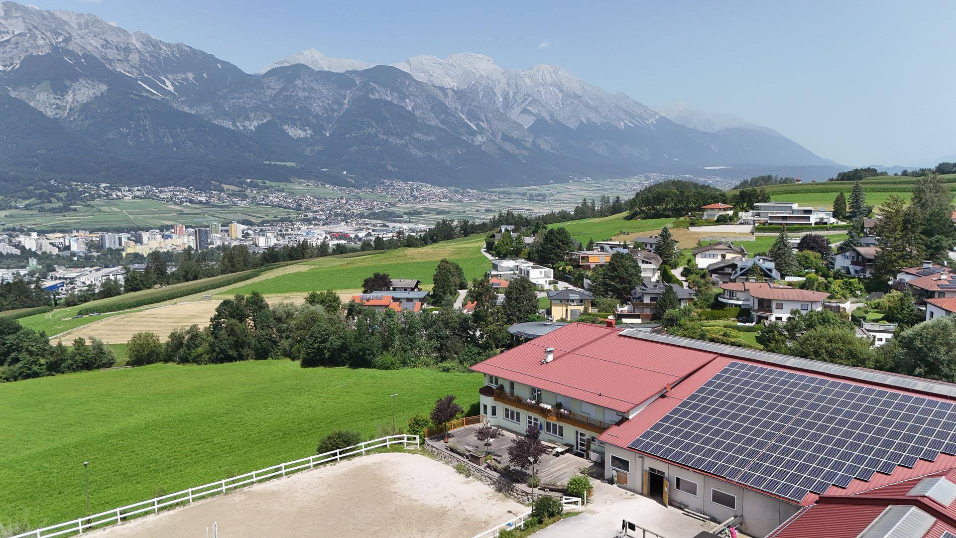 Luftaufnahme des Anwesens mit Reitplatz, Stallungen und Wohngebäude vor einer weiten Berg- und Stadtlandschaft.