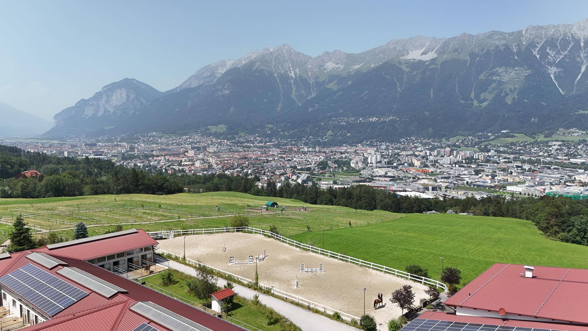 Weitläufige Luftaufnahme des Reitplatzes und der Gebäude mit Blick auf die Stadt und die umliegenden Berge.