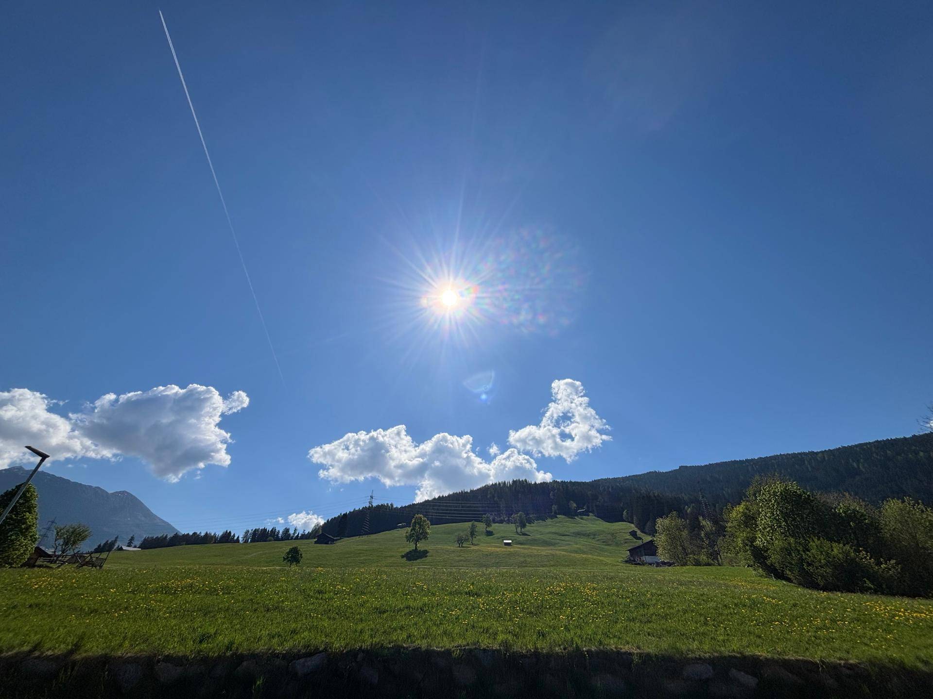 Malerische Landschaft mit grünen Hügeln, vereinzelten Bäumen und strahlendem Sonnenschein am blauen Himmel.