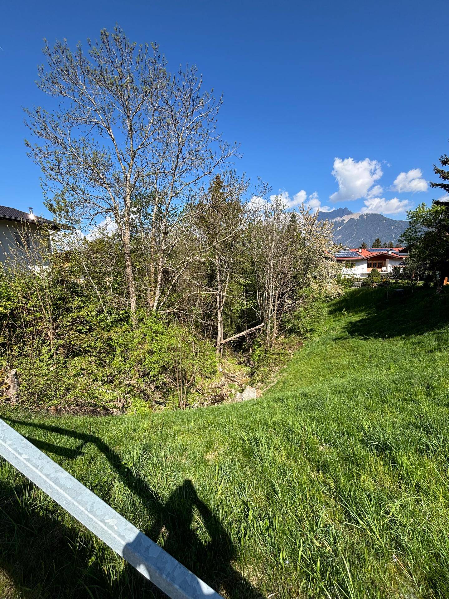 Grüne Böschung führt zu einem bewaldeten Bereich mit Bach und Bergblick im Hintergrund unter blauem Himmel.