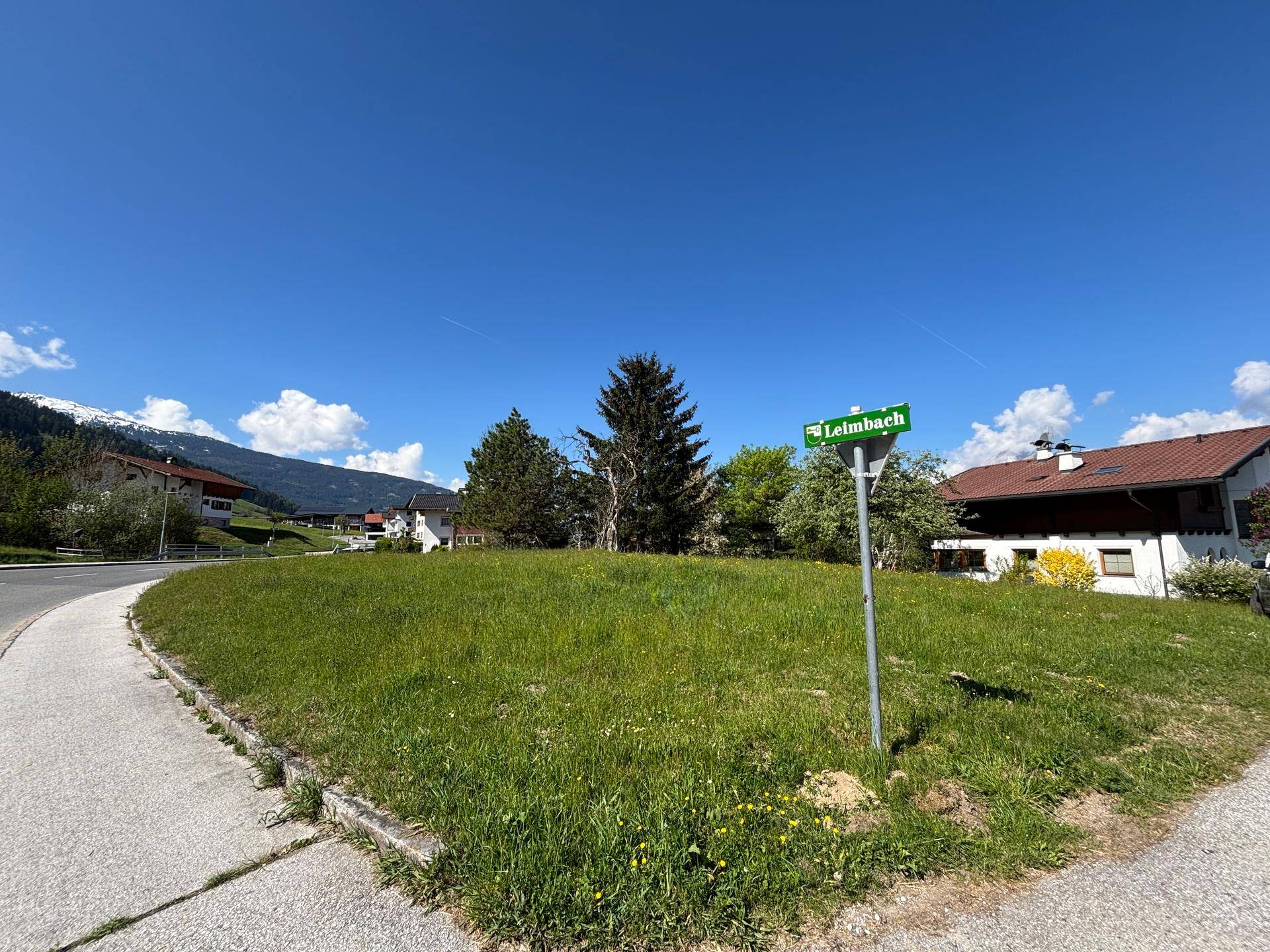 Grüne Parzelle an einer Straßenkreuzung mit Straßenschild und Bergblick im Hintergrund unter blauem Himmel.