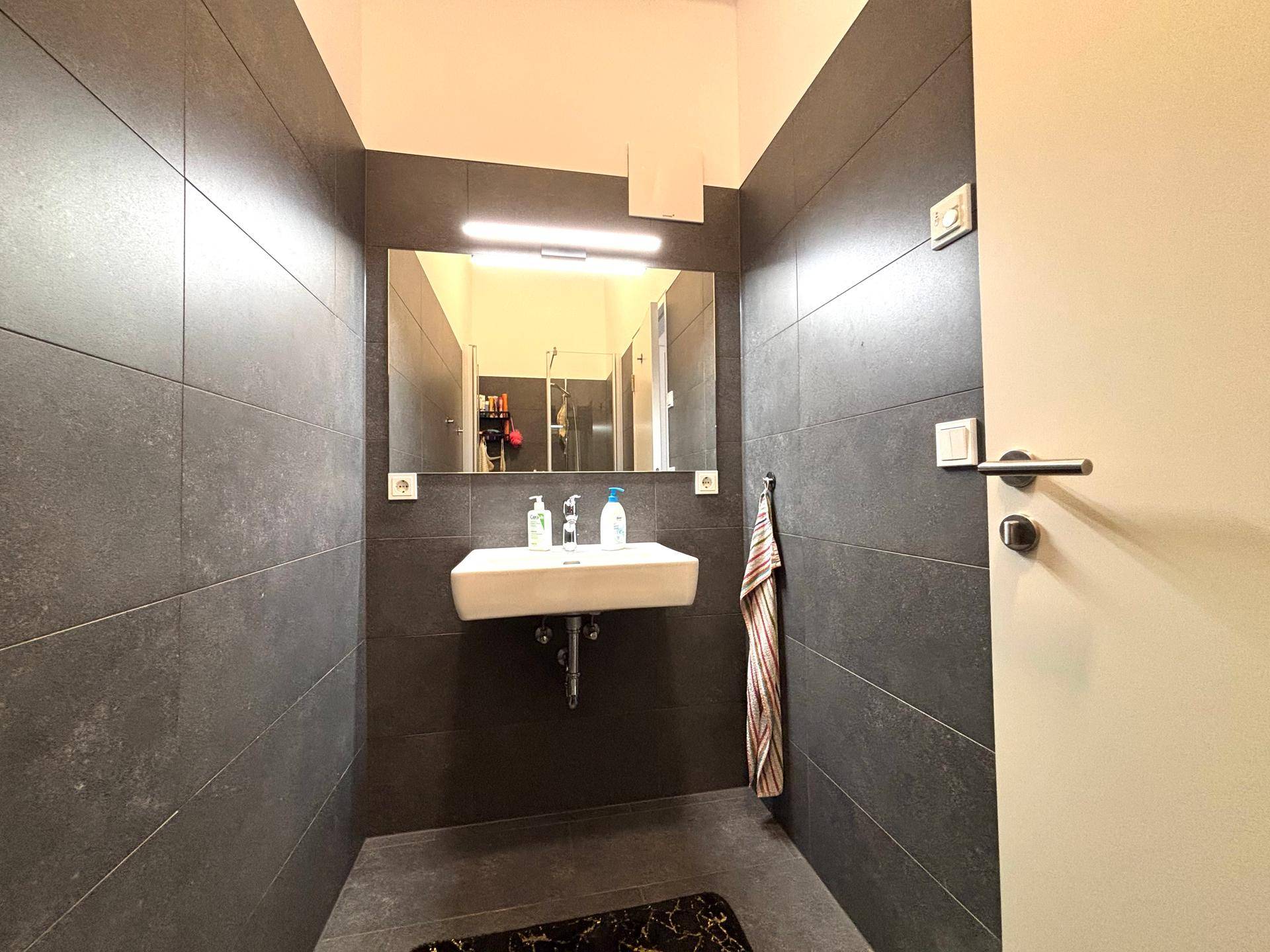 Badezimmer mit modernem Waschbecken, Spiegel und dunklen großformatigen Fliesen.