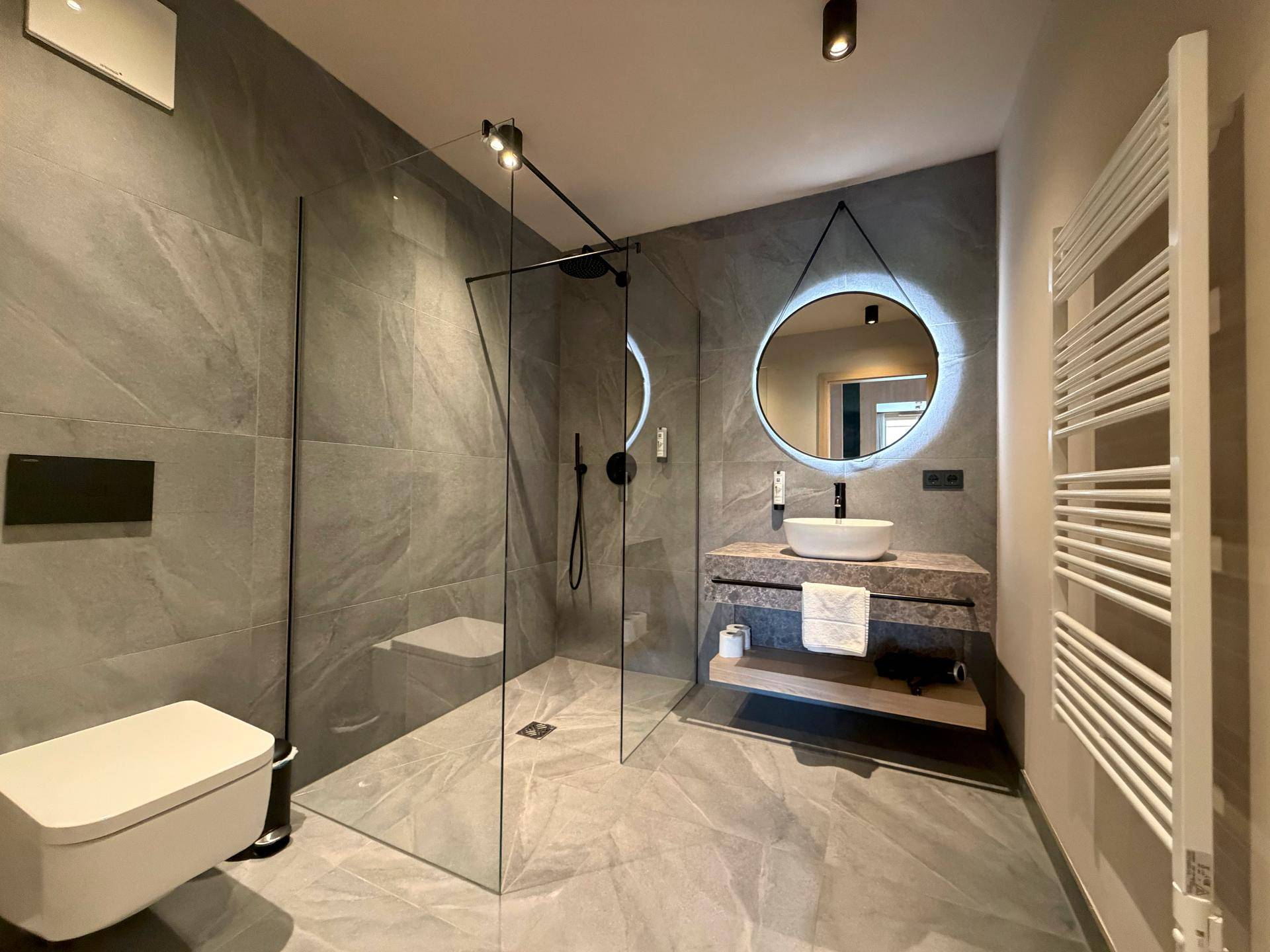 Modernes Badezimmer mit grauen Fliesen, begehbarer Dusche und beleuchtetem Spiegel.