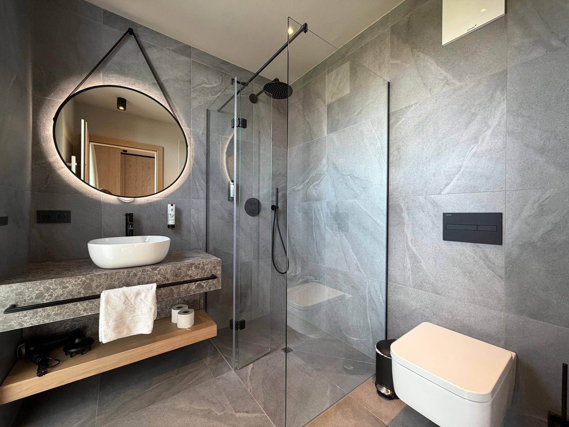 Modernes Badezimmer mit grauen Fliesen, begehbarer Dusche und beleuchtetem Spiegel.