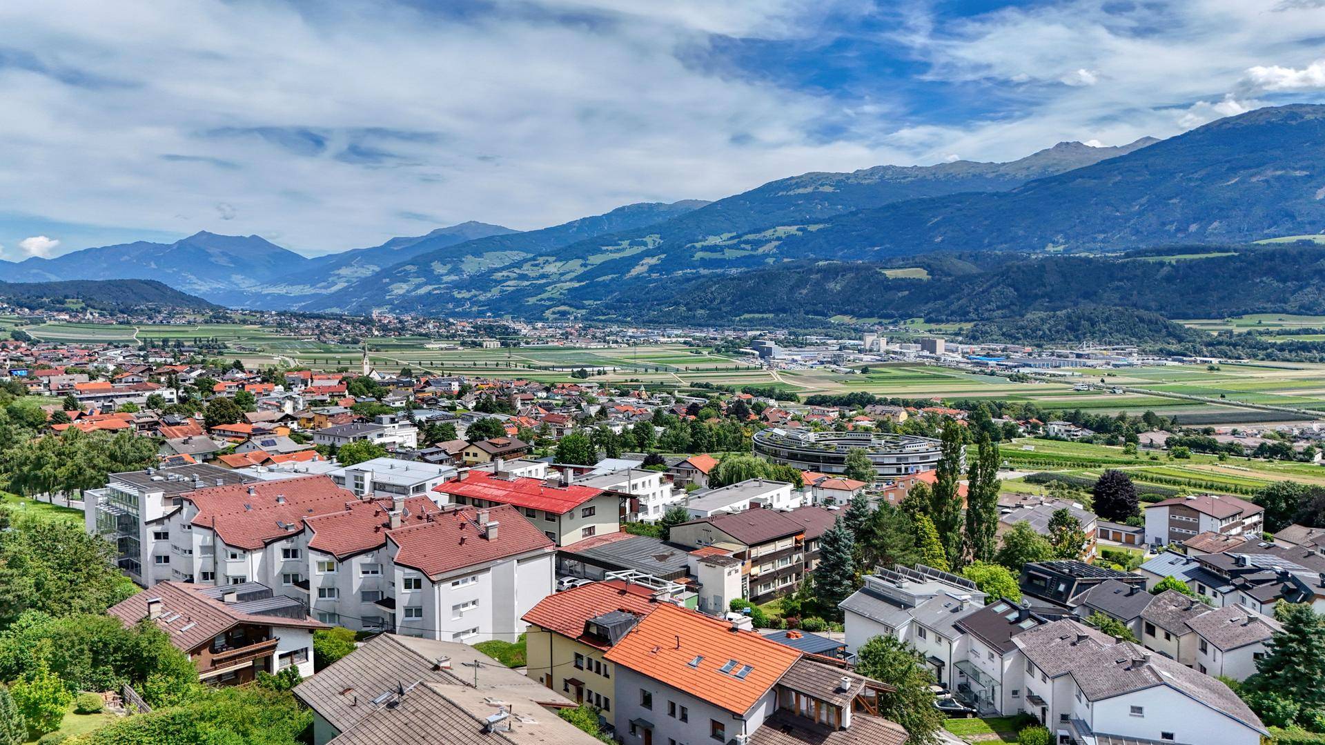 Luftaufnahme einer Wohnsiedlung mit zahlreichen Häusern, grünen Feldern und einem weiten Bergpanorama.