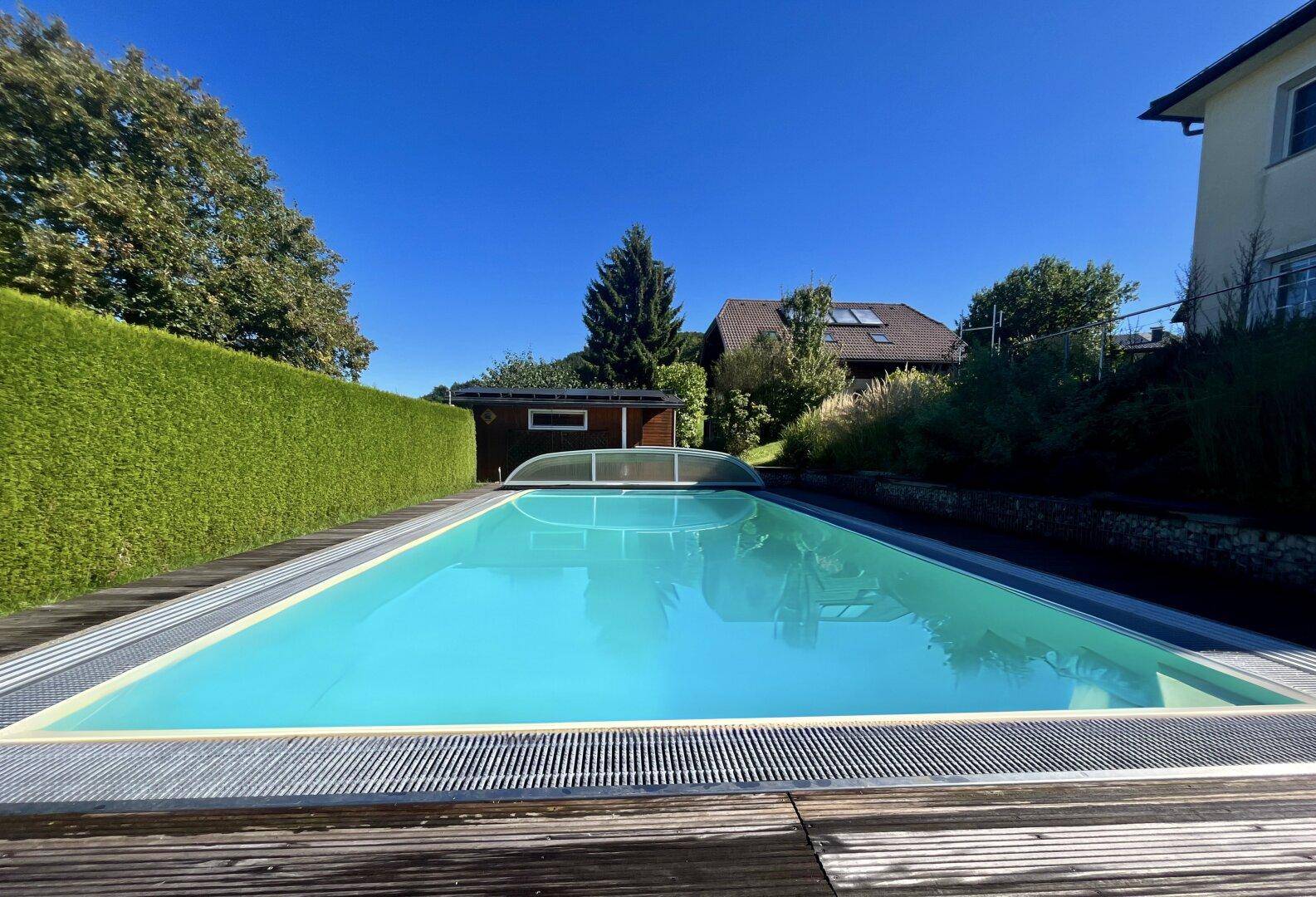 Außenpool mit Holzdeck und einer Abdeckung, umgeben von grüner Hecke.