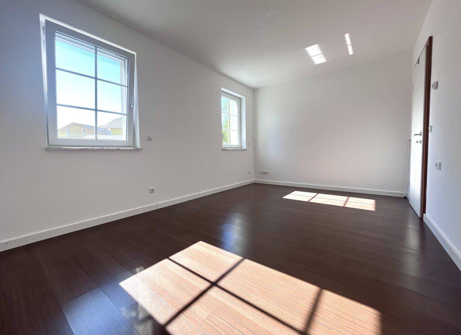 Helles Zimmer mit dunklem Holzboden und großen Fenstern, ideal als Schlaf- oder Arbeitszimmer.