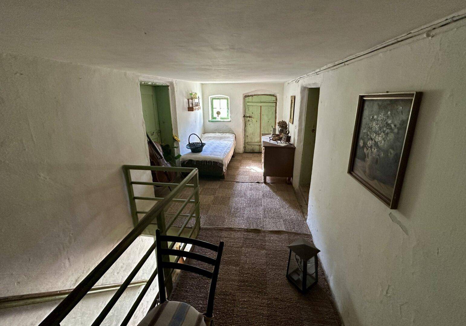 Heller Flur mit einem Einzelbett, kleinem Fenster und rustikalem Geländer, einfach eingerichtet.