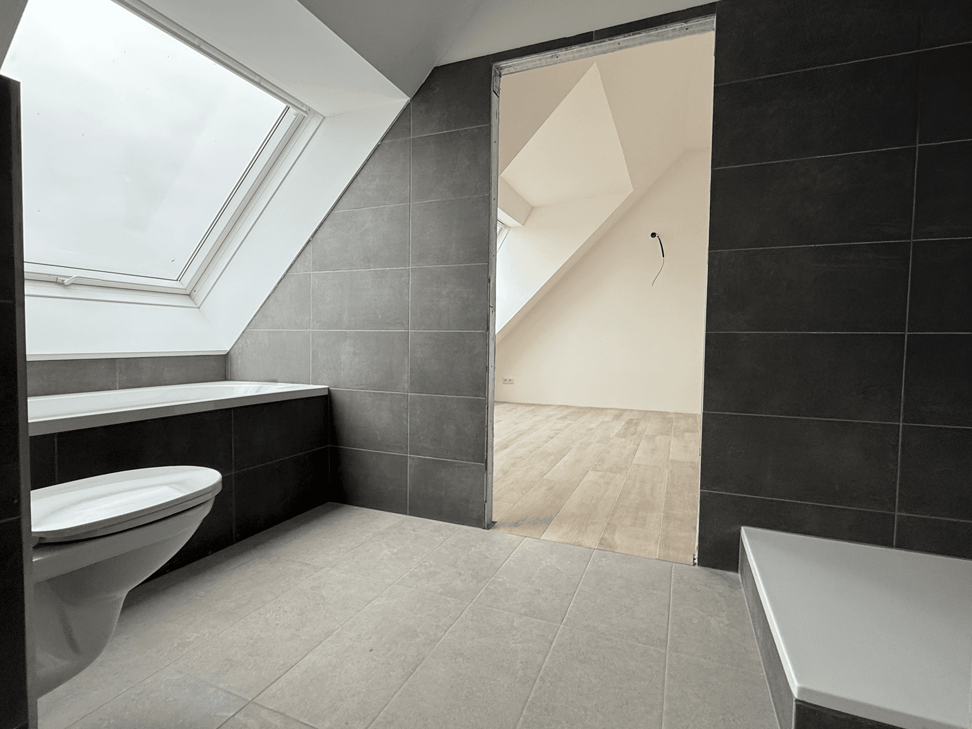 Modernes Badezimmer mit Badewanne, dunklen Fliesen, Wand-WC und Dachfenster.