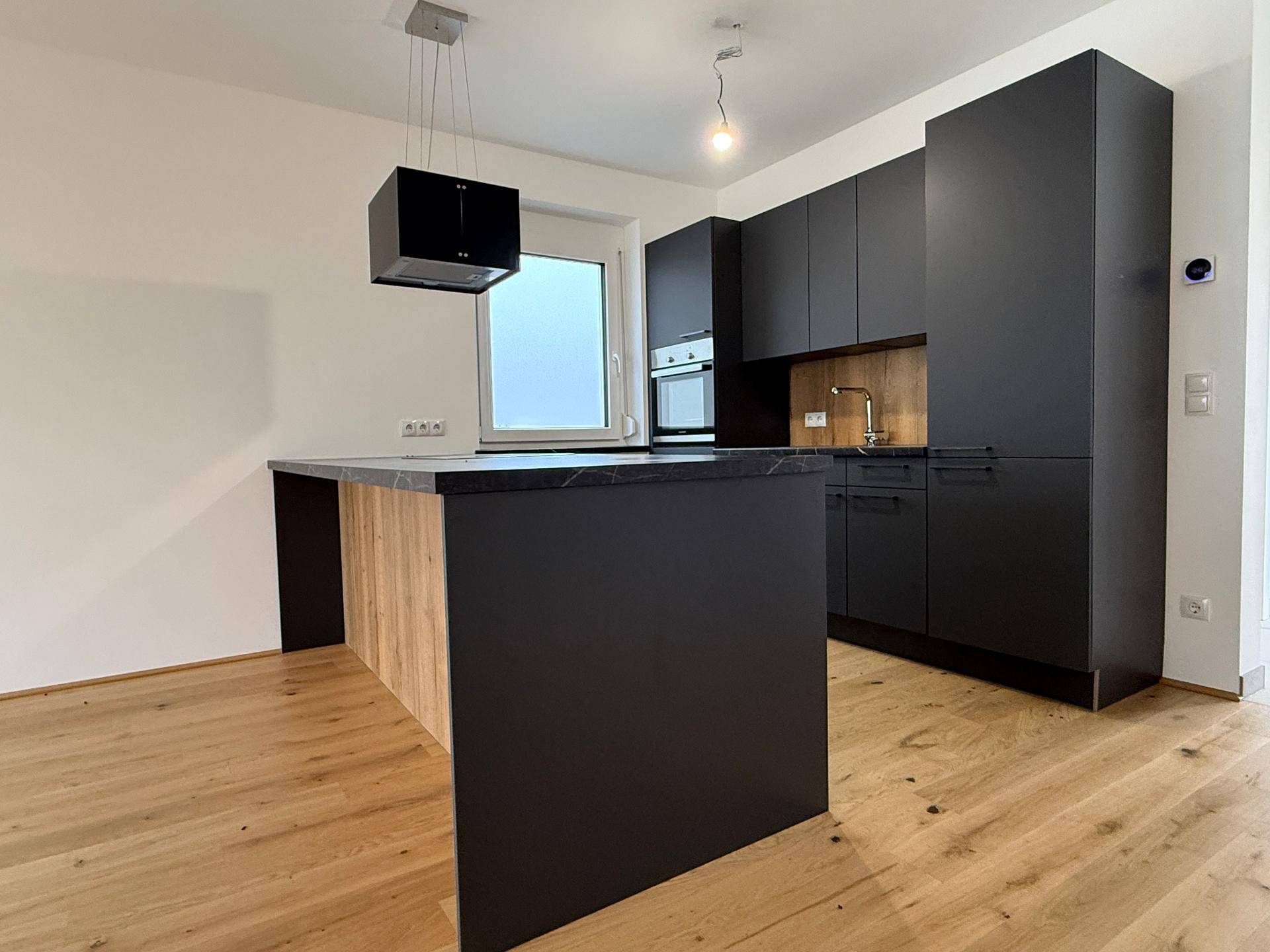 Moderne schwarze Einbauküche mit Kücheninsel, Holzdetails und Parkettboden in hellem Raum.