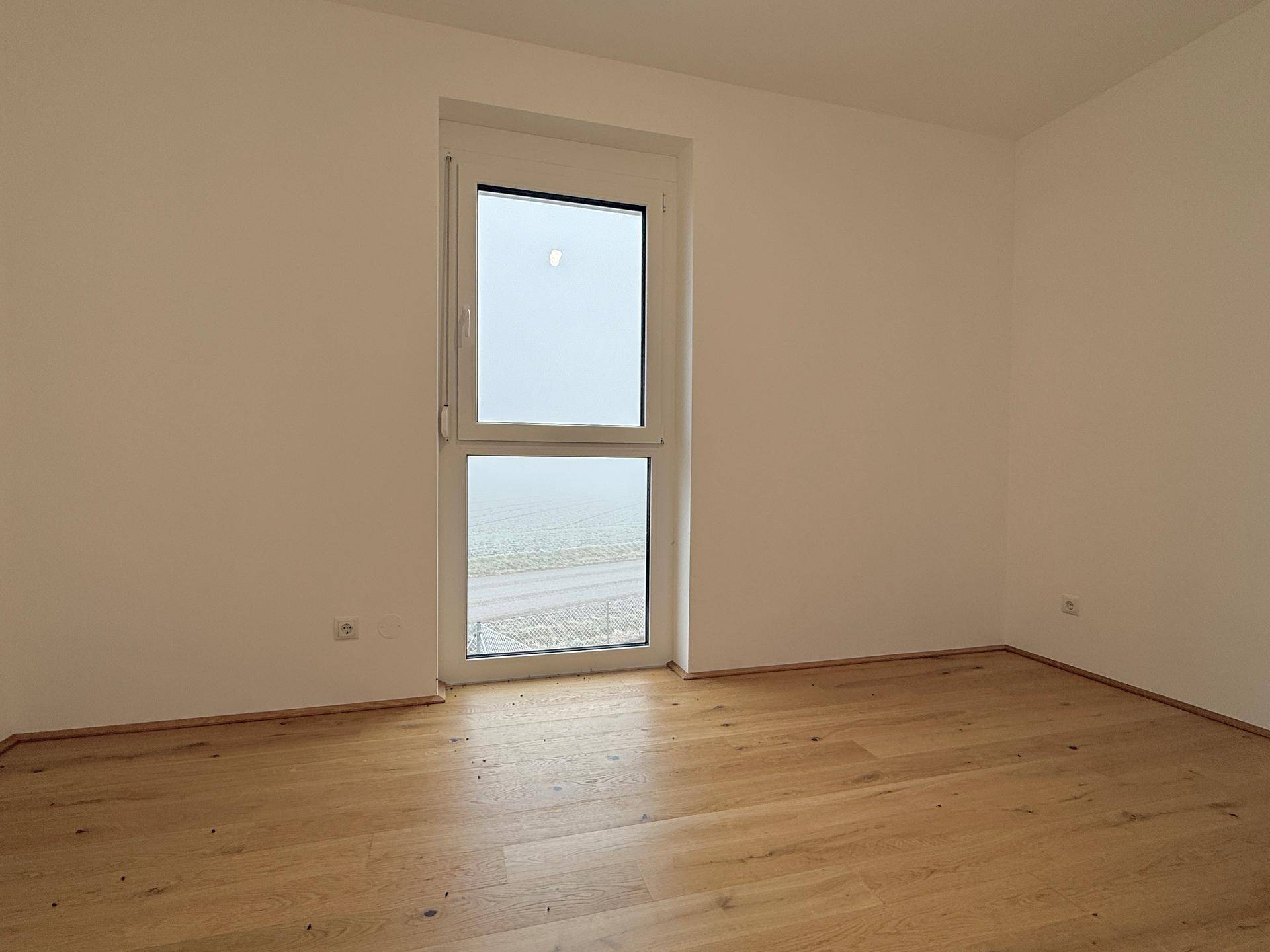 Leeres Schlafzimmer mit Holzboden und großem Fenster, das viel Tageslicht hereinlässt.