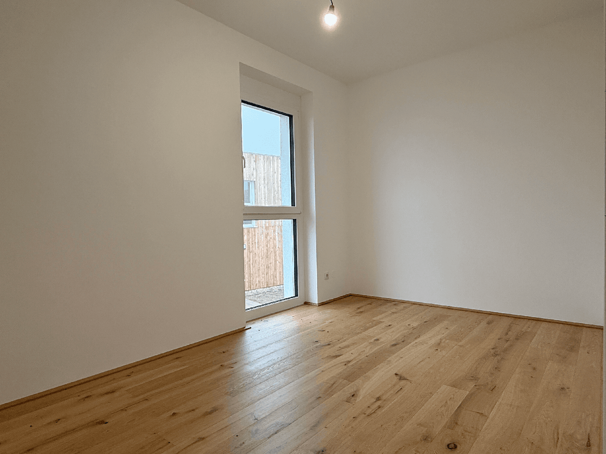 Leeres Zimmer mit Holzboden und großem Fenster, ideal als Schlaf- oder Arbeitszimmer.
