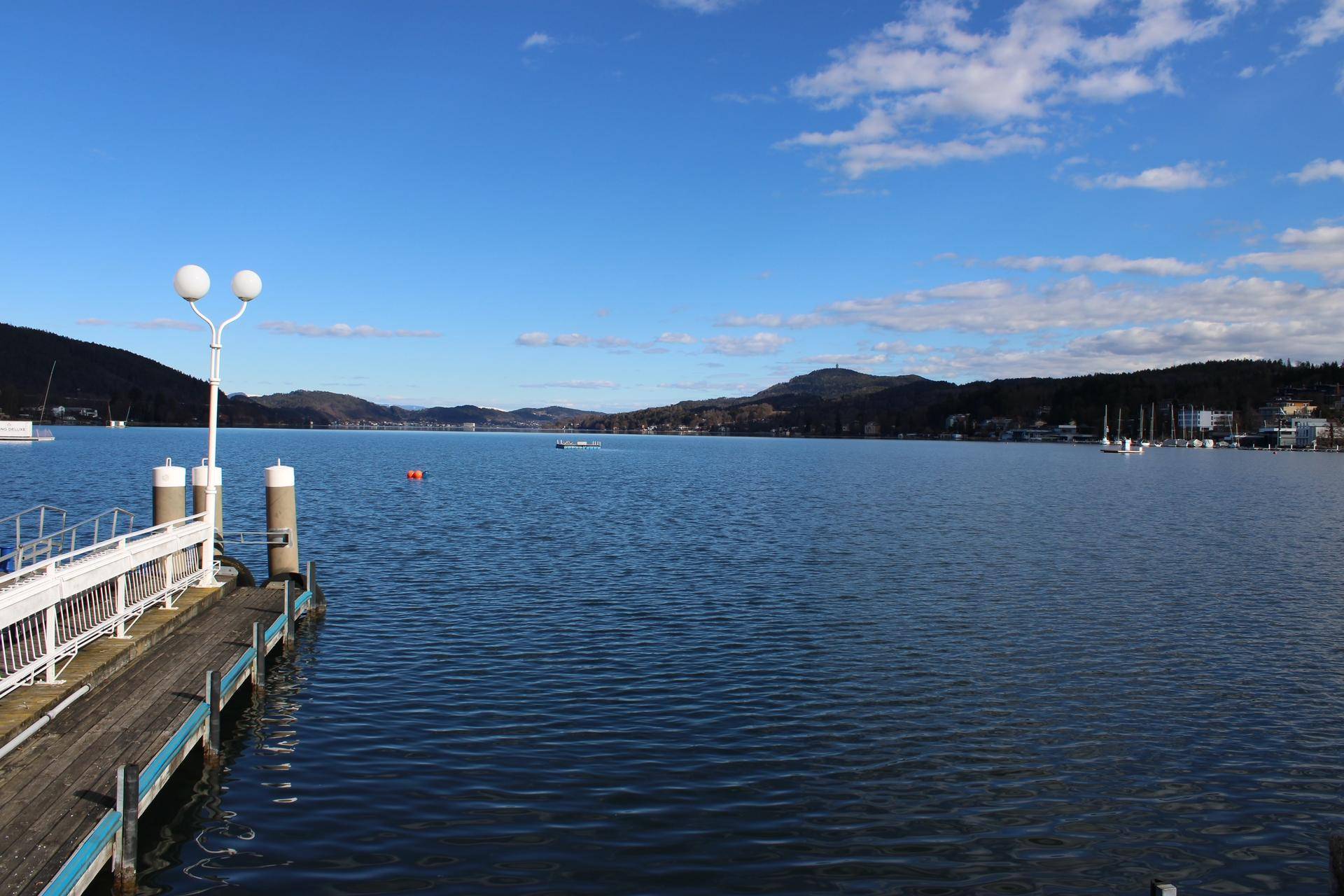 Weitläufiger Seeblick mit einem Holzsteg und Bergen im Hintergrund unter blauem Himmel.