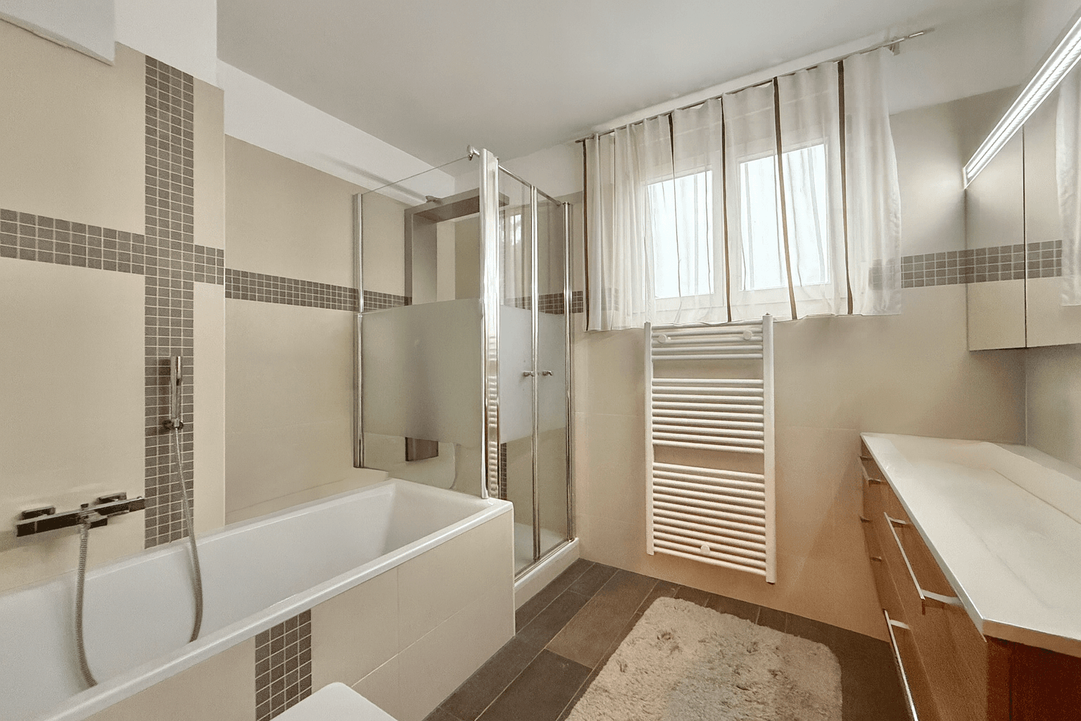 Helles Badezimmer mit Badewanne, Duschkabine, Fenster und einem praktischen beheizten Handtuchhalter.