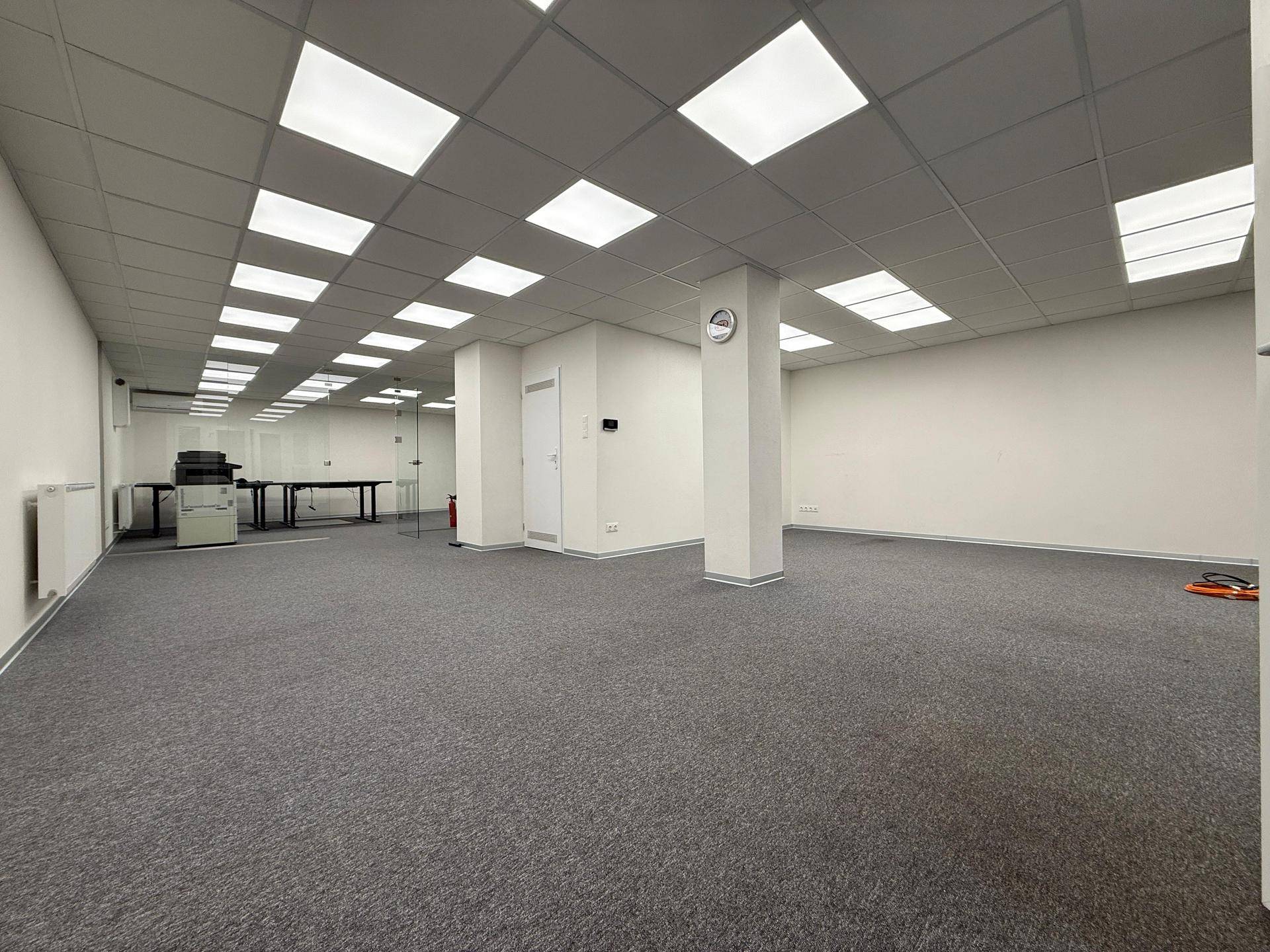 Weitläufiges Büro mit Teppichboden, Säulen und abgehängter Decke, bietet viel Platz für verschiedene Arbeitsbereiche.