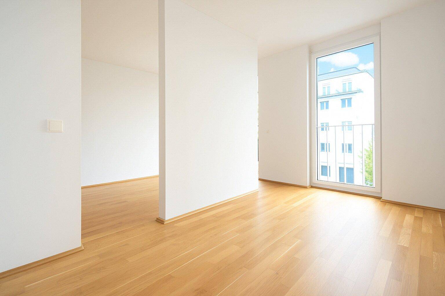 Lichtdurchfluteter Raum mit Holzboden und Fenster mit Blick auf ein gegenüberliegendes Gebäude.