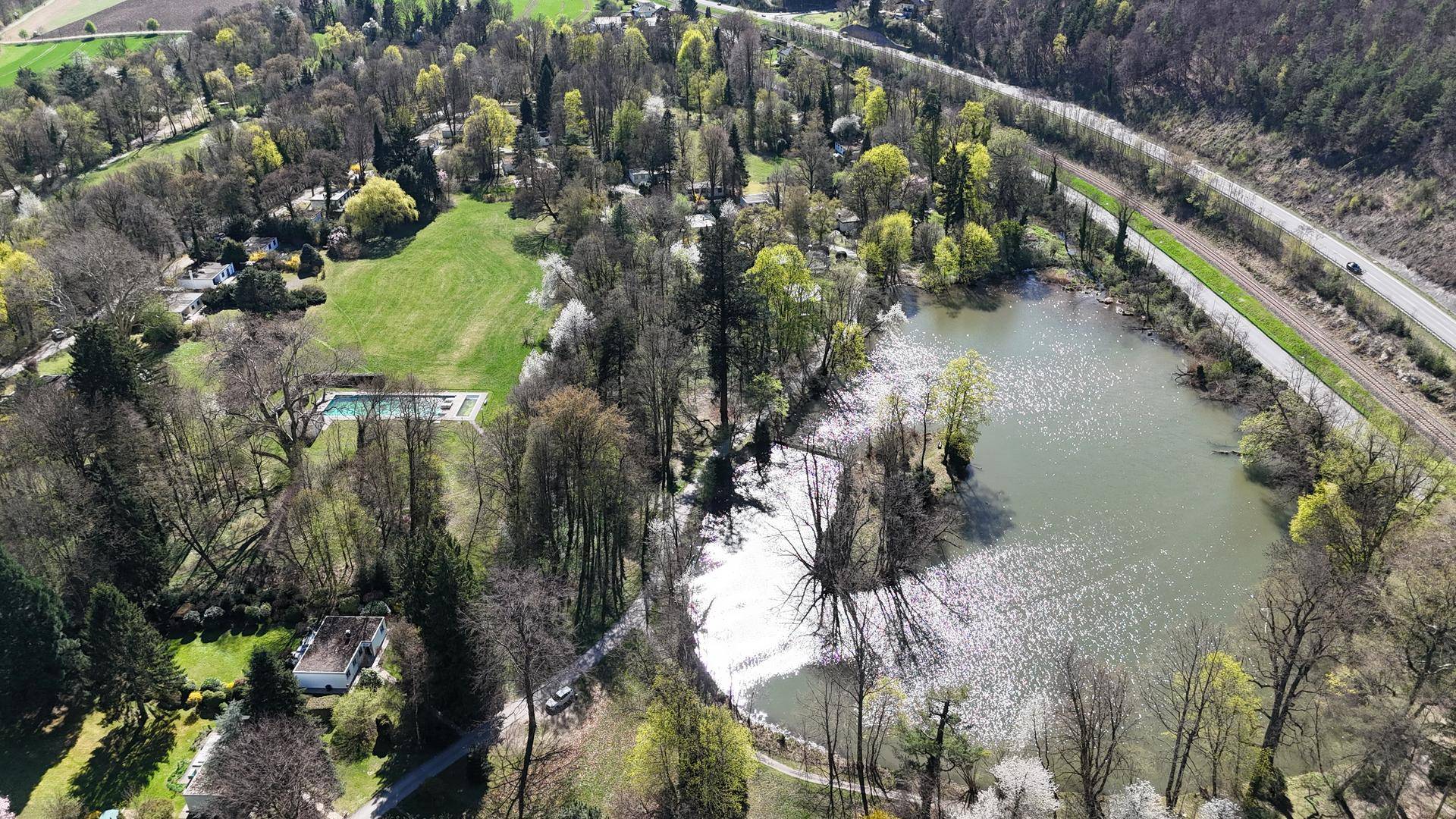 Weitläufige Luftaufnahme mit See, Straße und ausgedehnten Grünflächen, inklusive des Pools der Anlage.