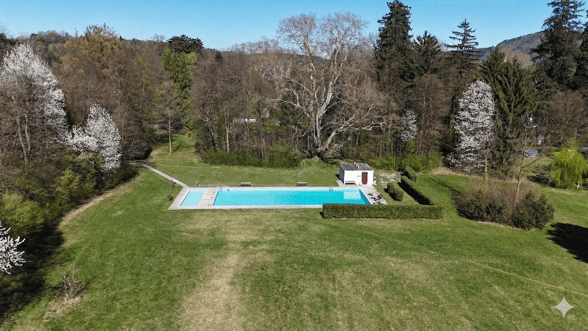 Großer Außenpool in einer weitläufigen Grünanlage, umgeben von Bäumen und einem kleinen Poolhaus.