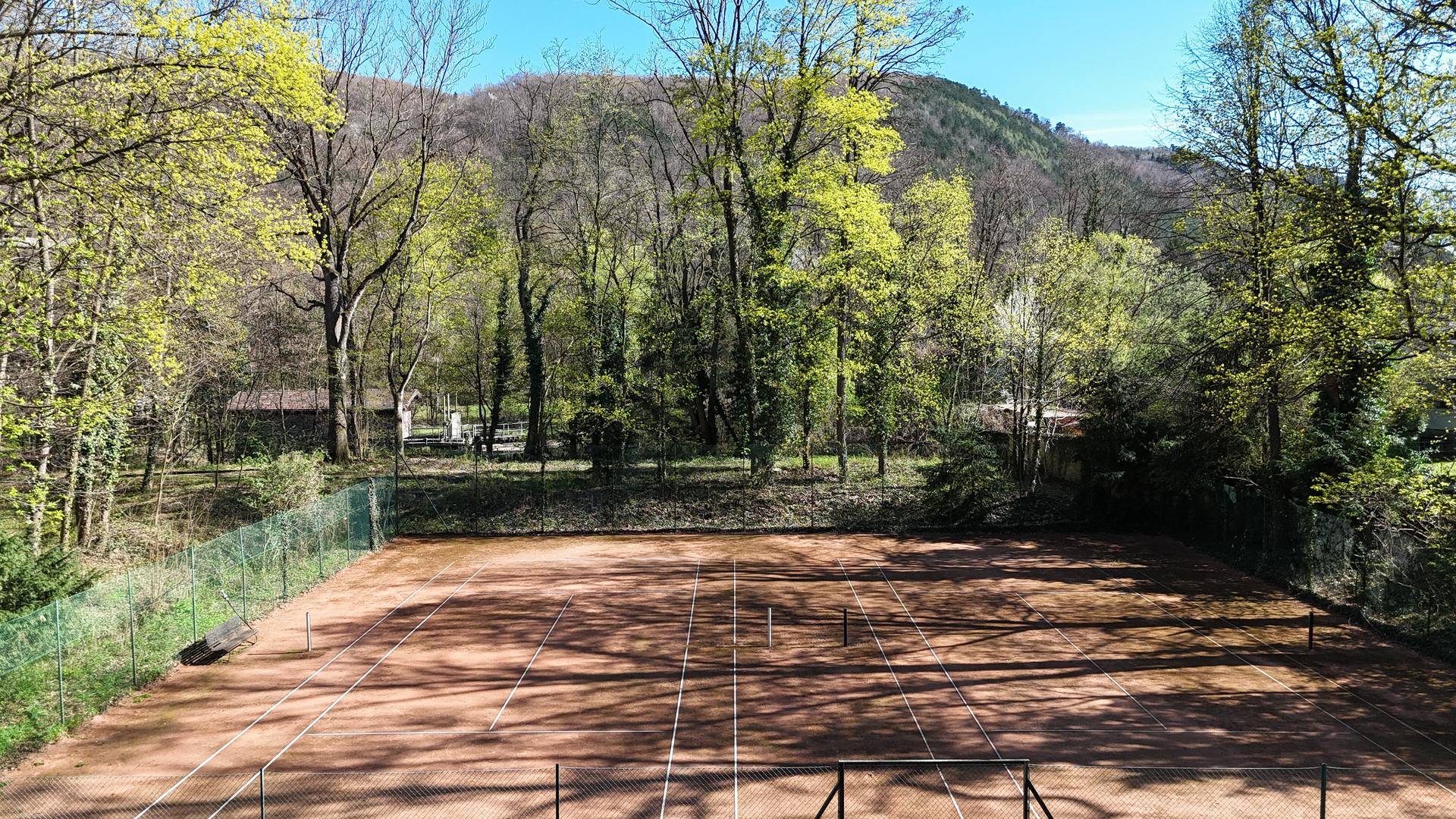 Außen-Tennisplatz umgeben von hohen Bäumen, bereit für sportliche Aktivitäten im Freien.